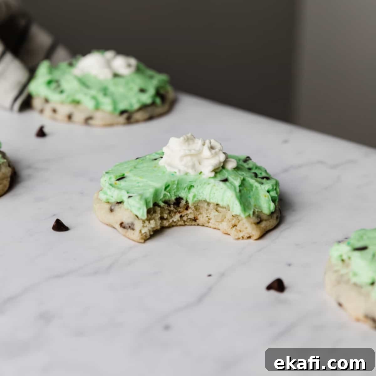 Ένα δαγκωμένο αντιγραφές Crumbl Mint Chip Ice Cream Cookie με πράσινο γλάσο μέντας και ένα κουταλάκι σαντιγί.