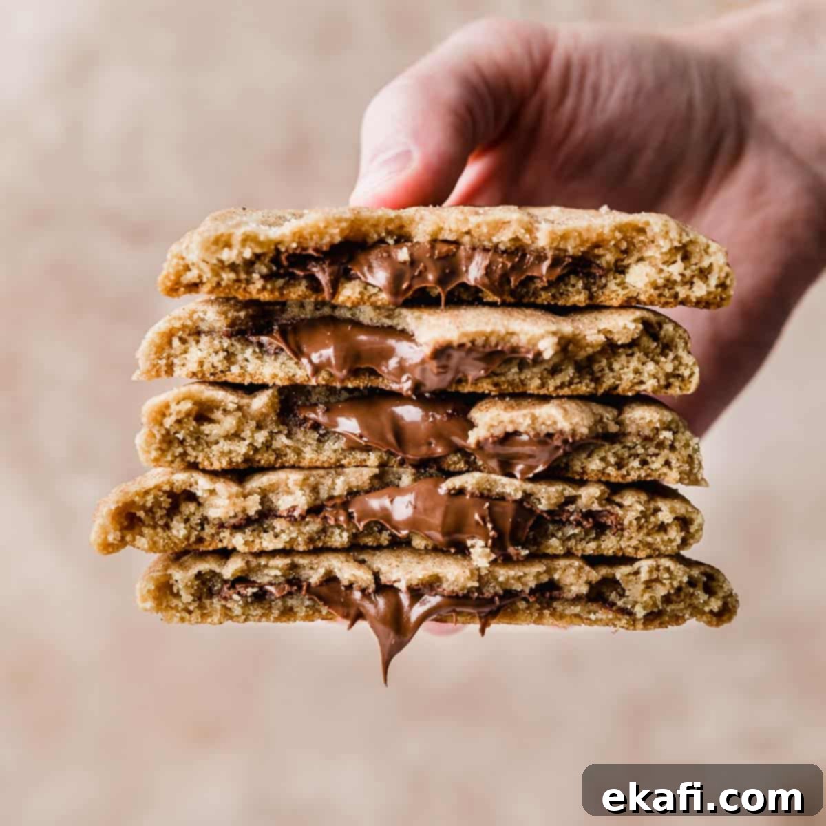 Ένα χέρι κρατάει μια στοίβα από 5 Crumbl Hazelnut Churro Cookies κομμένα στη μέση, σε μπεζ φόντο.