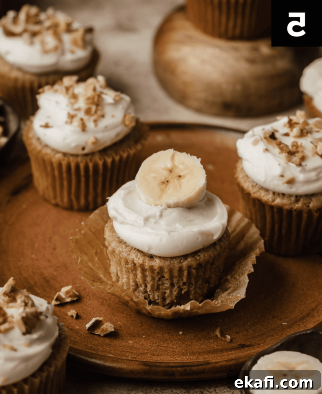 Συνταγή για Κάπκεϊκς Μπανάνας 5 Ένα cupcake μπανάνας γαρνιρισμένο με λευκό γλάσο τυριού κρέμας και μια φέτα φρέσκιας μπανάνας στην κορυφή, το cupcake βρίσκεται σε ένα καφέ πιάτο.