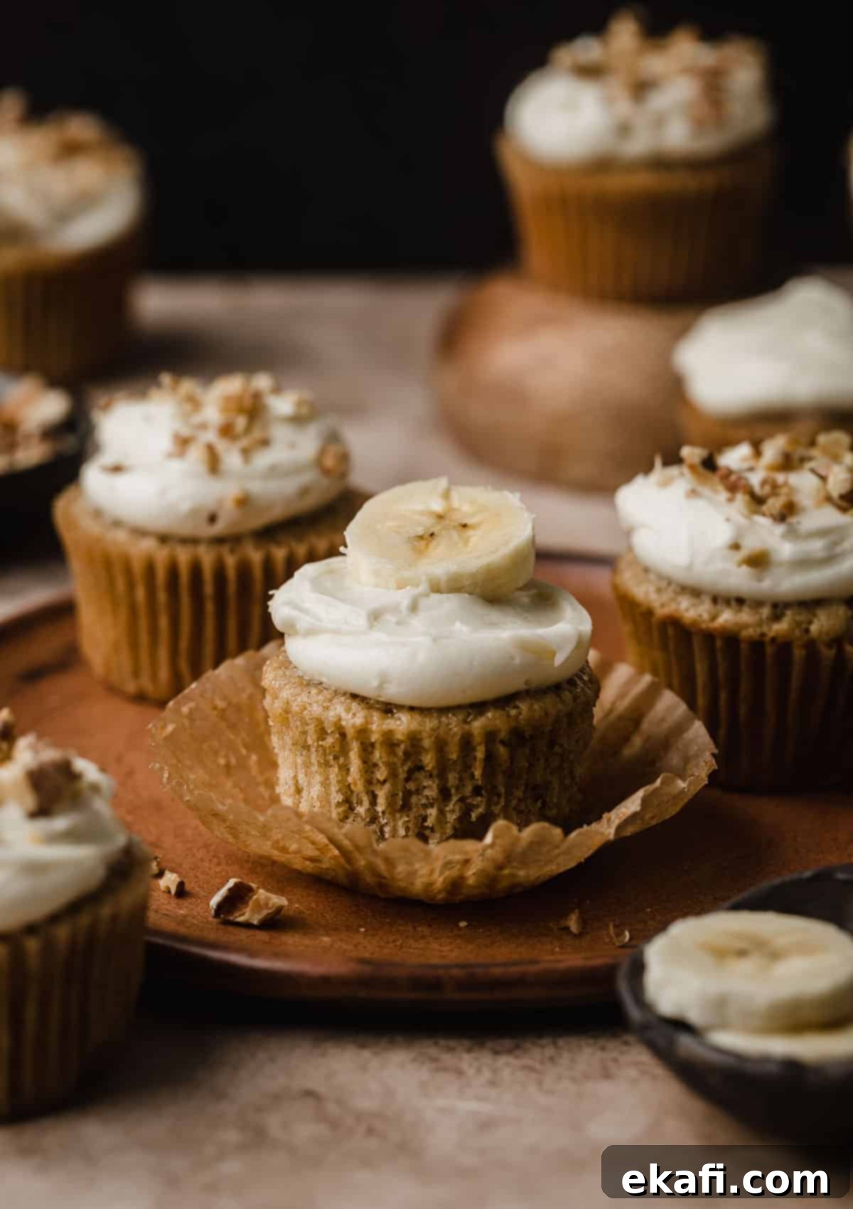 Συνταγή για Κάπκεϊκς Μπανάνας 7 Cupcakes μπανάνας σε ένα καφέ πιάτο με σκούρο καφέ φόντο, με κάποια από τα cupcakes γαρνιρισμένα με ψιλοκομμένα καρύδια.