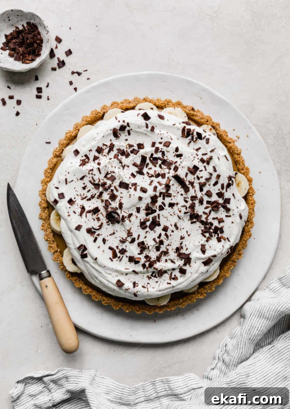Μια Banoffee Pie με σαντιγί και ξύσματα σοκολάτας σε ένα λευκό στρογγυλό πιάτο.