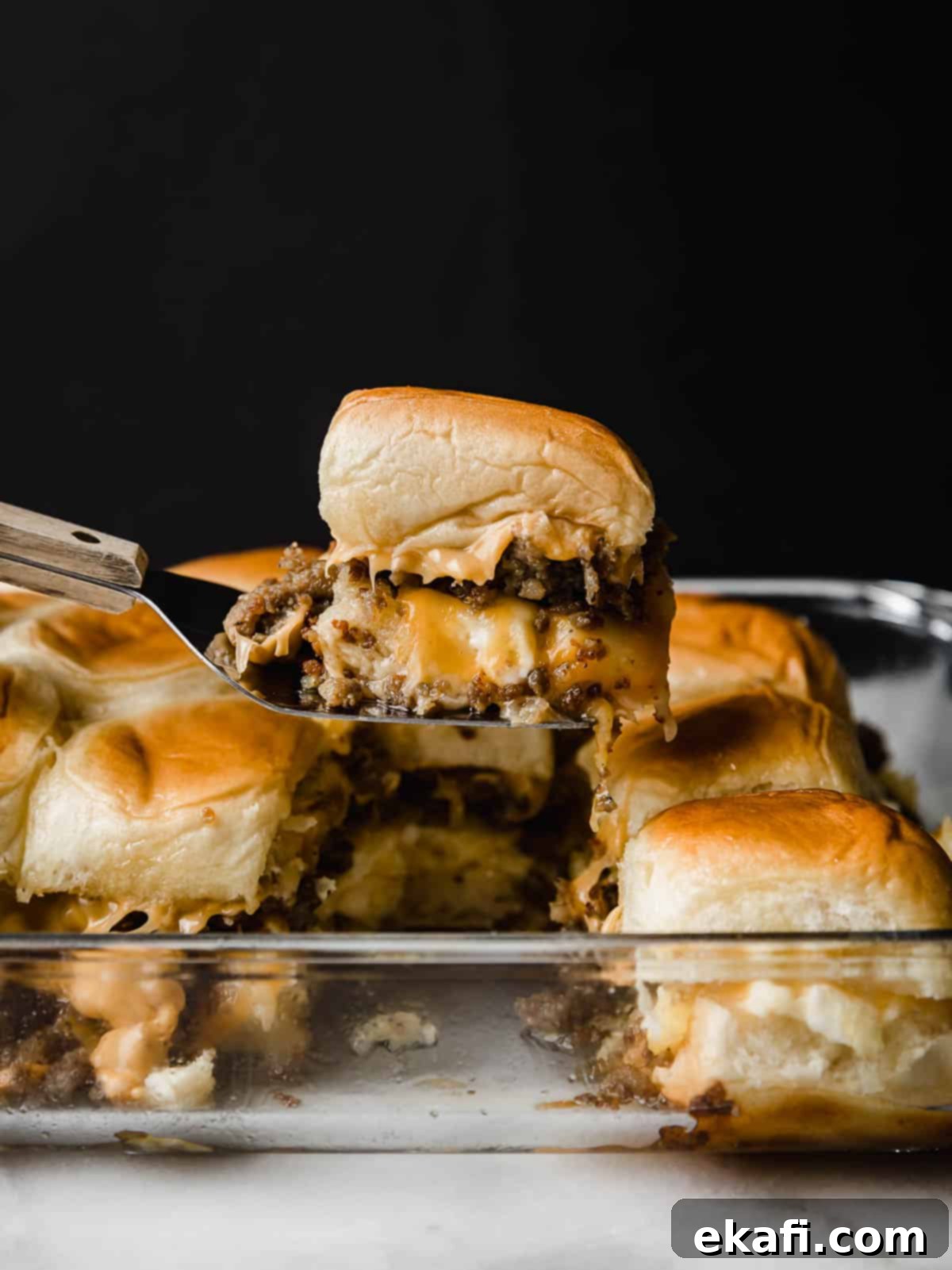 Μια συνταγή για Hawaiian Roll Breakfast Sliders κρατιέται με μια μεταλλική σπάτουλα μπροστά σε μαύρο φόντο.