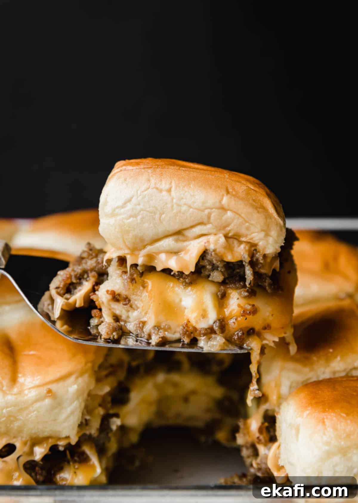 Μίνι Σάντουιτς Πρωινού 8 Εύκολα Breakfast Sliders με λουκάνικο και τυρένια ομελέτα φτιαγμένα σε ψωμάκια Hawaiian, ισορροπώντας σε μια μεταλλική σπάτουλα μπροστά σε μαύρο φόντο.