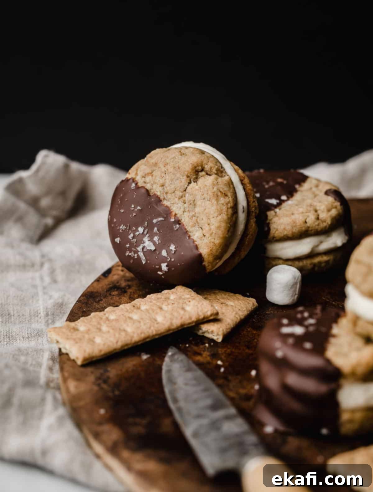 Μπισκότα Σάντουιτς Σ'μόρες 7 Ένα μπισκότο s'mores σάντουιτς με κρέμα μαρσμέλοου και βουτηγμένο σε λιωμένη σοκολάτα, ακουμπισμένο σε καφέ επιφάνεια.