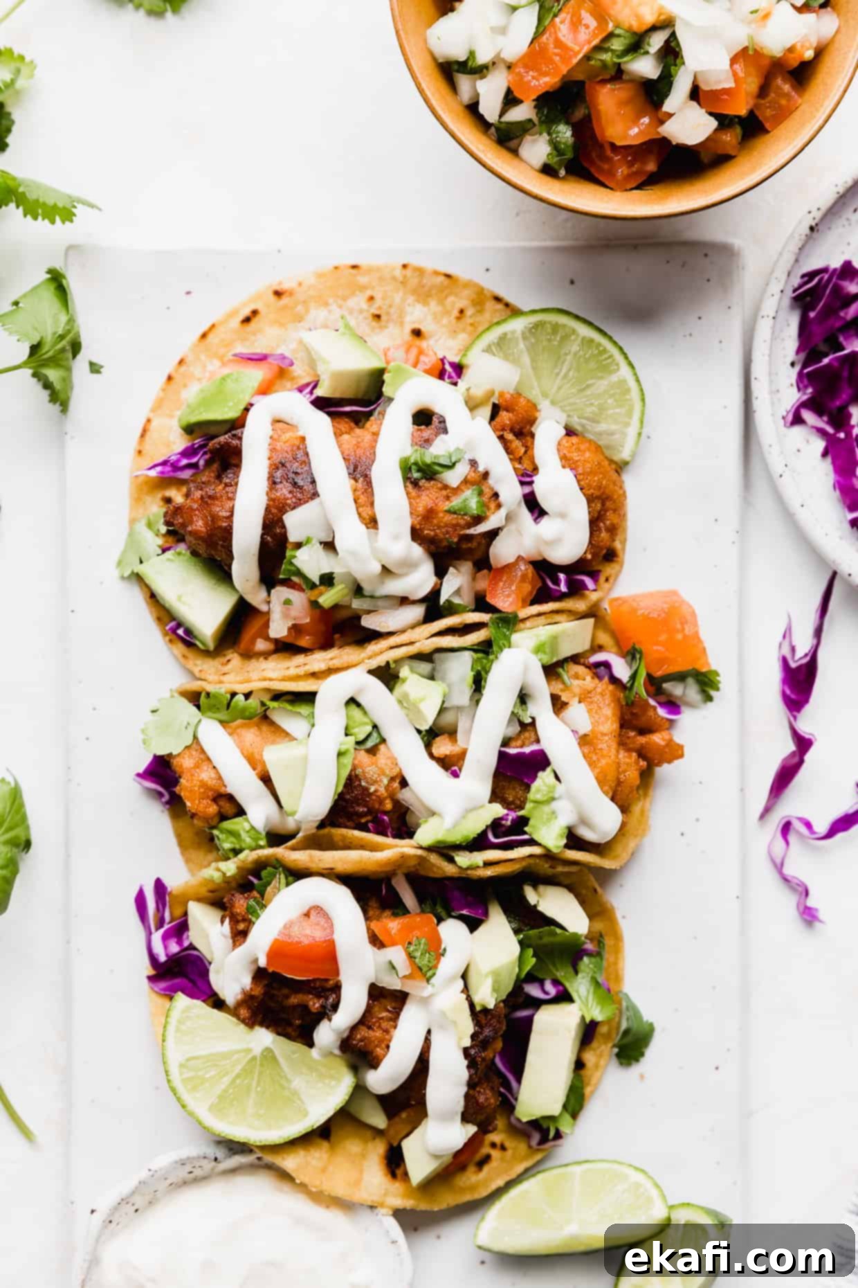 Crispy Fish Taco Bliss 11 Three Baja Fish Tacos elegantly arranged on a white plate, enhanced with fresh lime wedges, creamy avocado slices, a drizzle of white sauce, and colorful shredded cabbage.