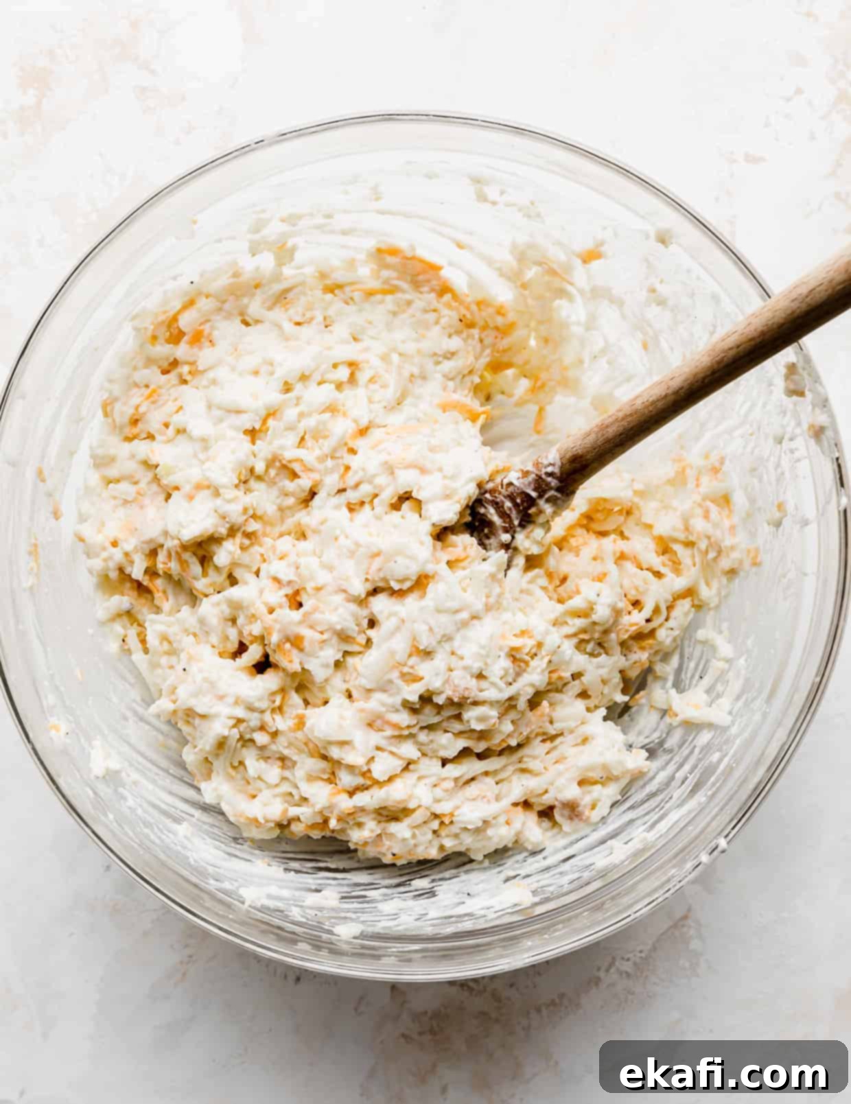Soul-Soothing Spuds 5 A large glass bowl with all funeral potatoes ingredients mixed together by a wooden spoon, creating a creamy potato mixture.