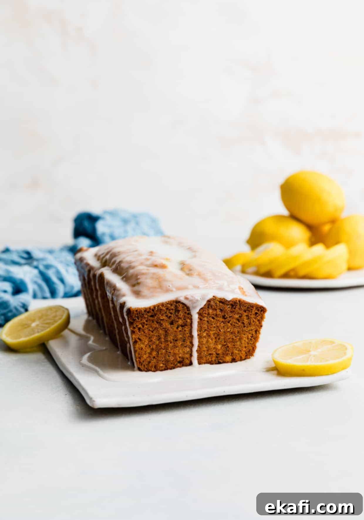 Cake Mix Lemon Poppy Seed Loaf 8 A loaf of Lemon Poppy Seed Bread drizzled with a white glaze against a white background, with yellow lemons stacked on a plate near the loaf.
