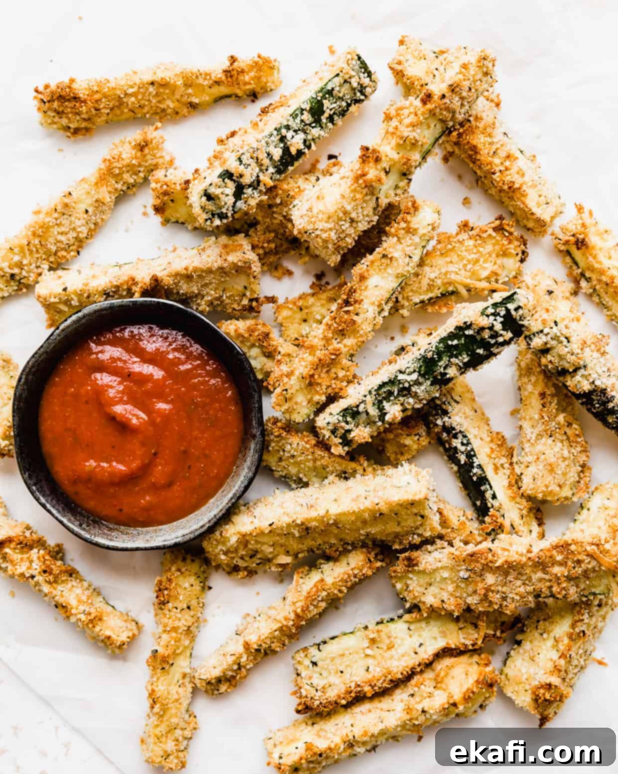 Crispy Baked Zucchini Fries 2 Baked Zucchini Fries on a white parchment paper with a small bowl of marinara sauce next to the fries.