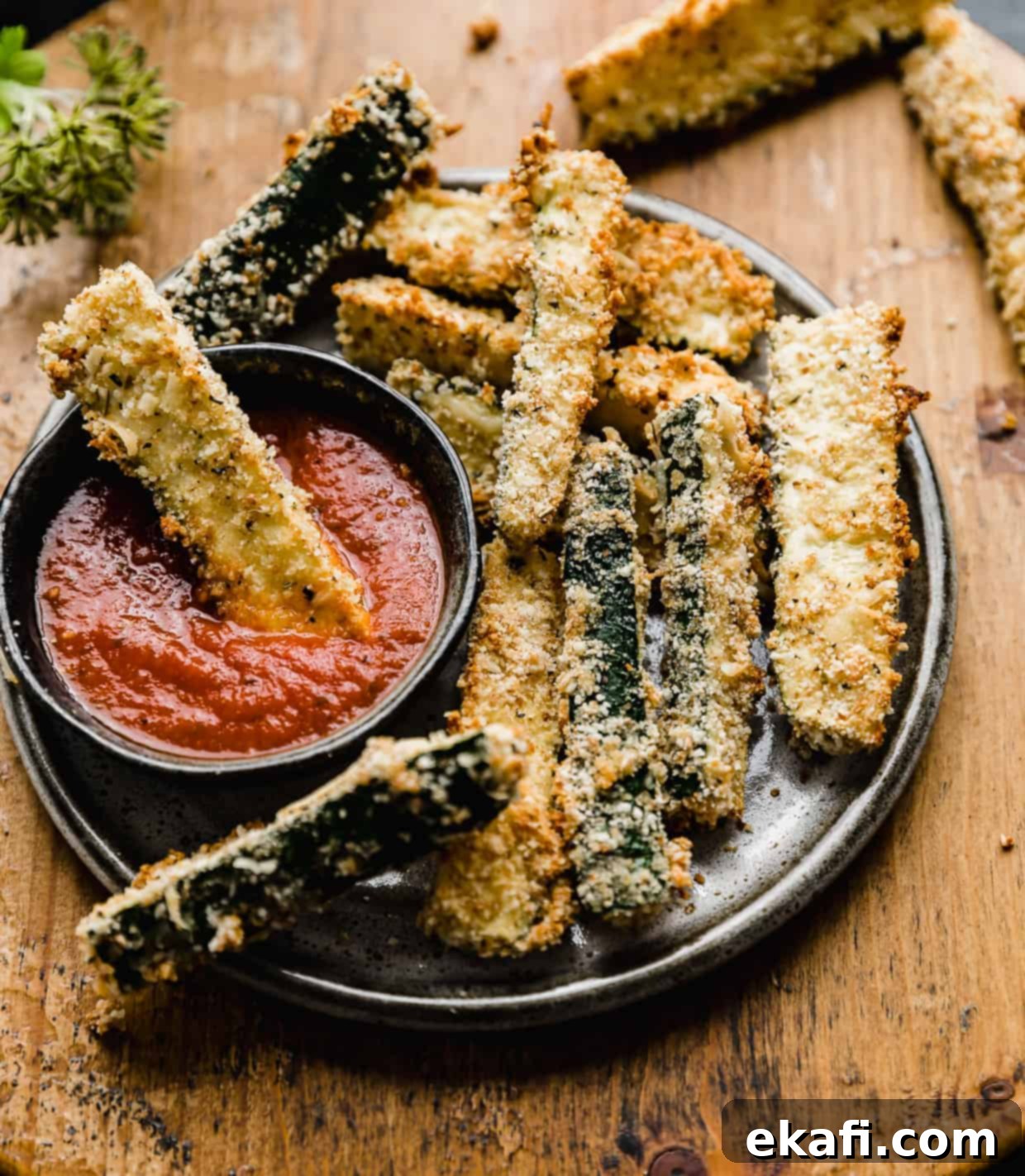 Crispy Baked Zucchini Fries 8 Zucchini fries on a black plate against a wooden background, with one zucchini fry dipped into pizza sauce.