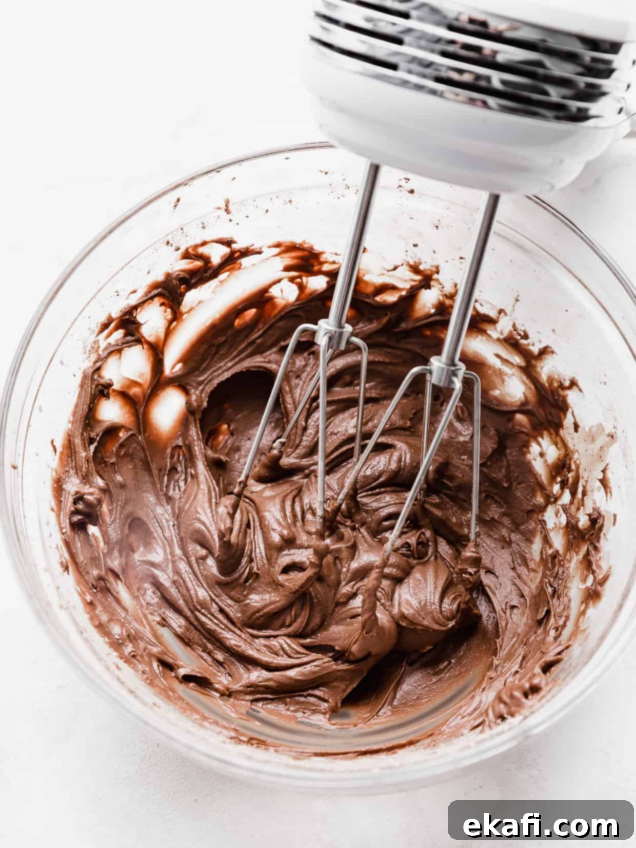 School Cafeteria Brownies 8 Hand beaters actively mixing rich chocolate brownie frosting in a clear glass bowl, showing its smooth texture.