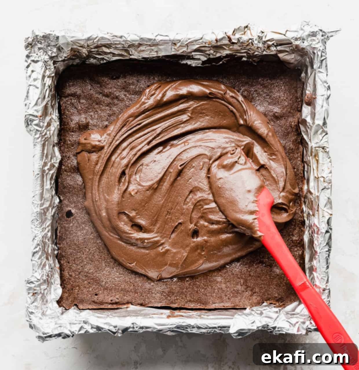 School Cafeteria Brownies 9 A red spatula gracefully spreading thick chocolate brownie frosting across a pan of warm brownies, creating a smooth, even layer.