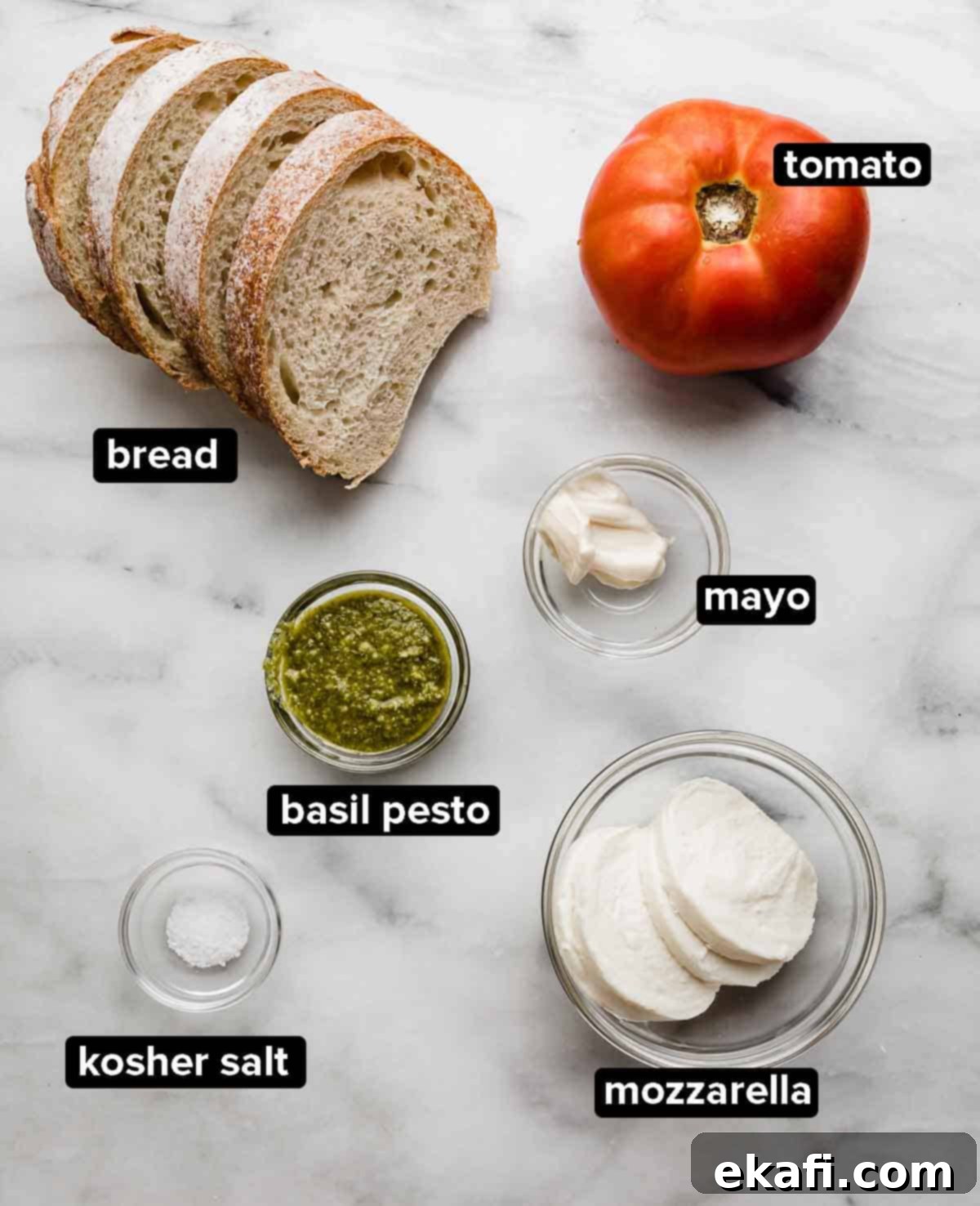 Pesto Mozzarella Sandwich 3 Pesto panini ingredients on a marble background, French bread, basil pesto, tomato, mayo, mozzarella, and salt.