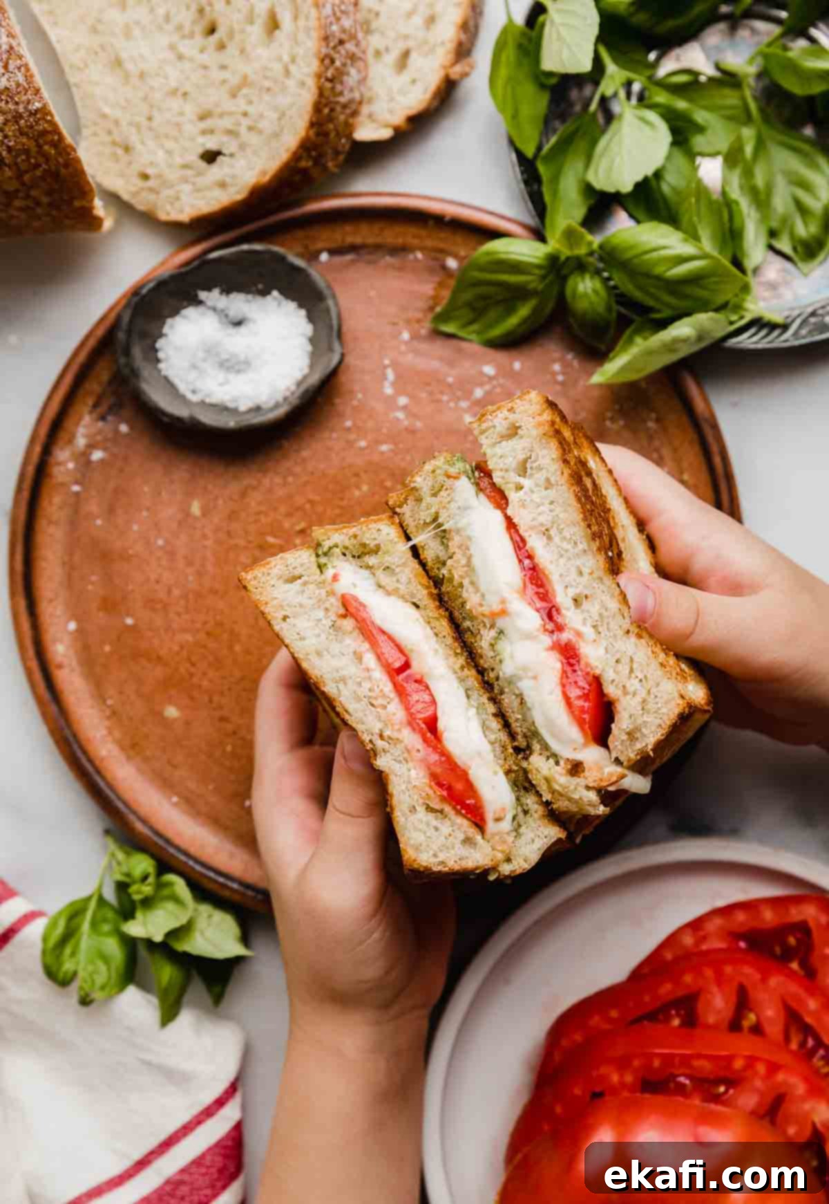 Pesto Mozzarella Sandwich 6 A pair of hands holding two halves of a Pesto Mozzarella Sandwich with tomato over a brown plate.