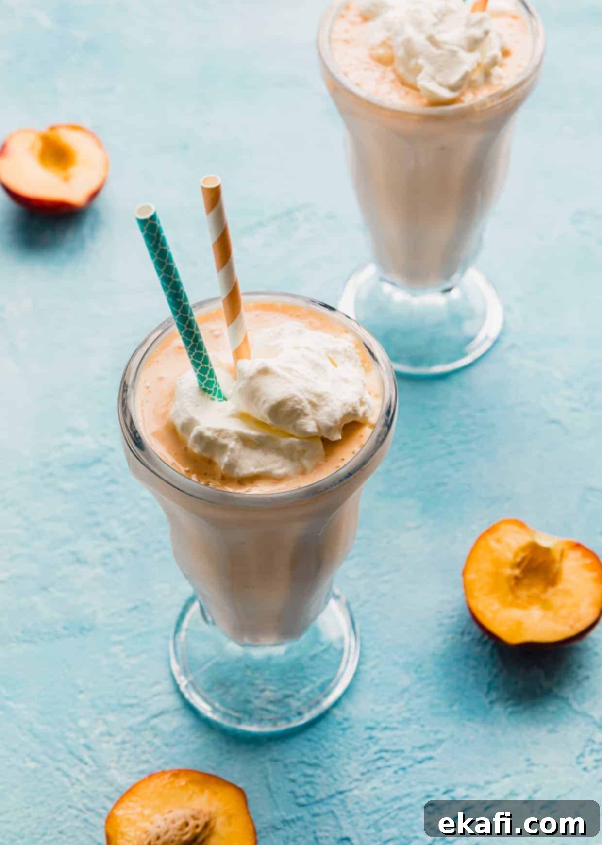 Velvet Peach Shake 7 A vibrant peach milkshake in a clear glass with blue and peach-colored straws, garnished with a peach slice, ready to be enjoyed.