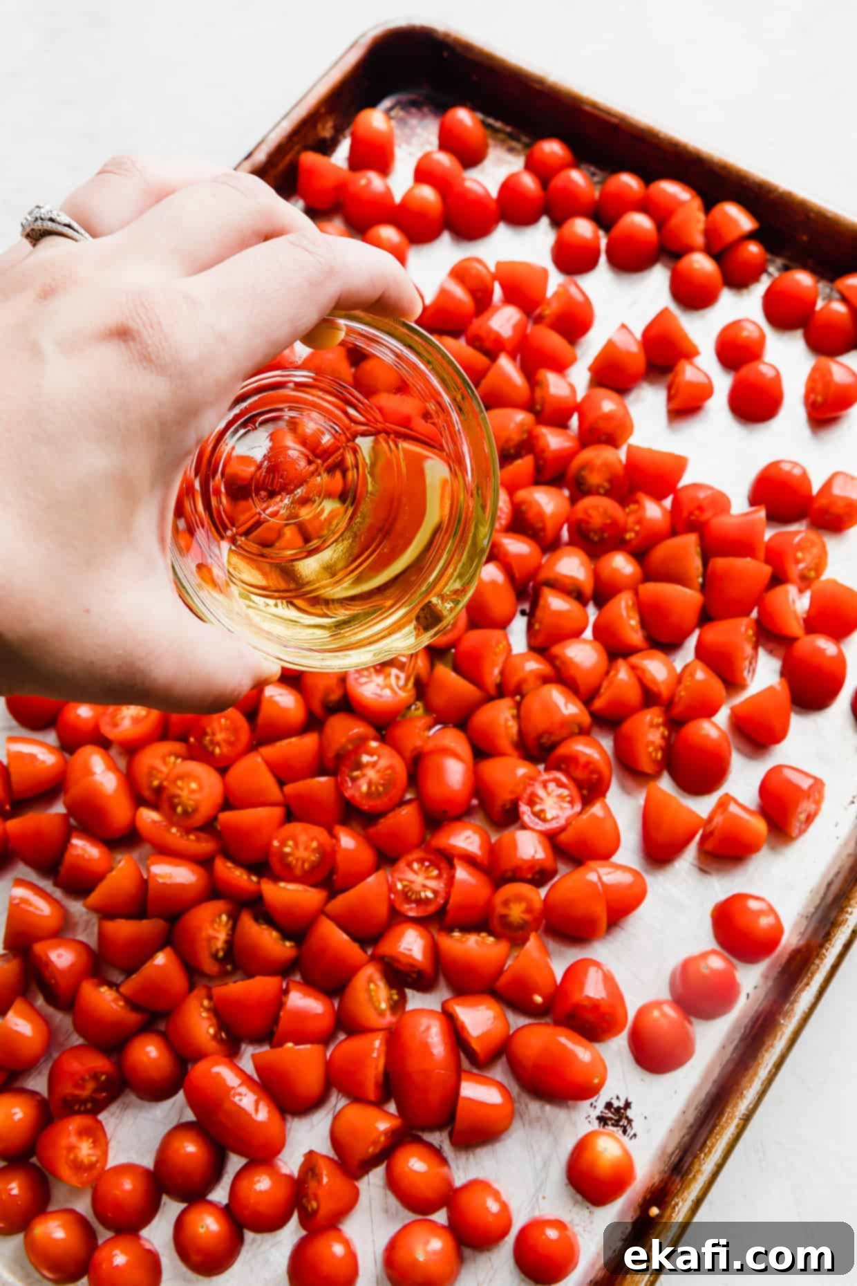 Blistered Grape Tomatoes 5 Olive oil being drizzled from a glass bowl over a generous amount of grape tomatoes on a baking sheet, ready for seasoning.