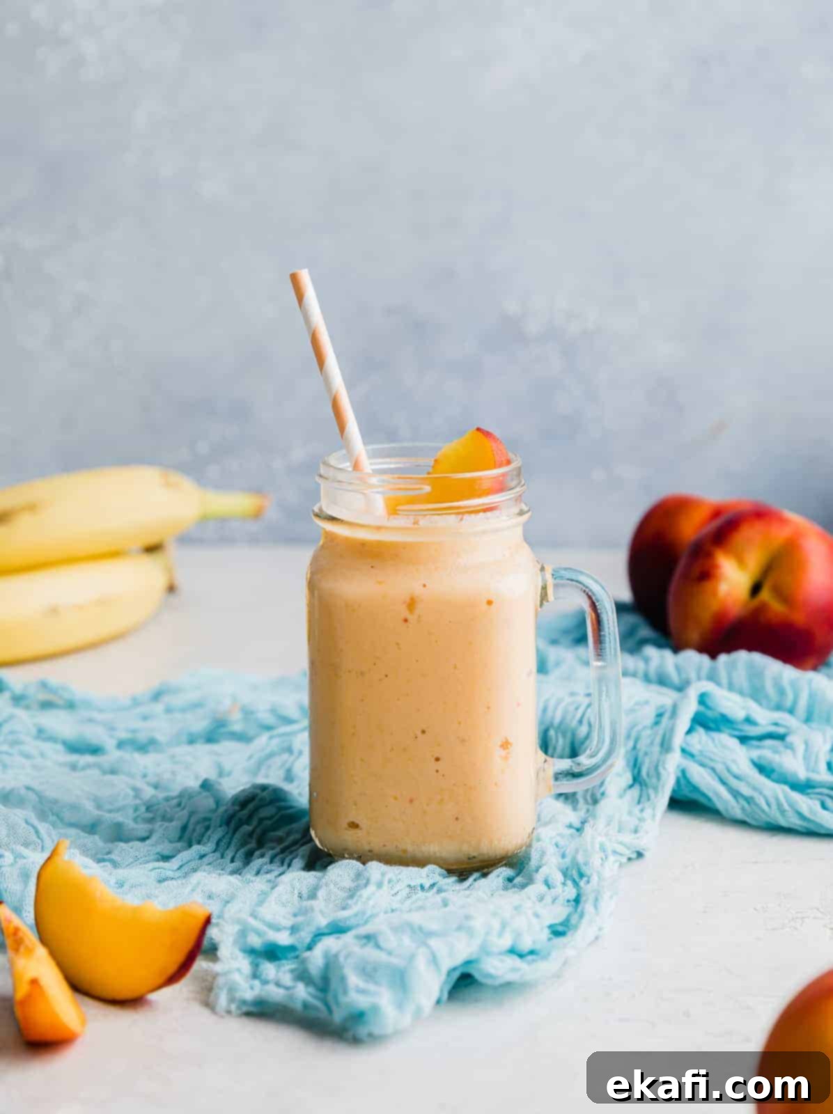 Creamy Peach Banana Bliss 7 A banana peach smoothie in a glass cup with fresh peaches and bananas in the background.