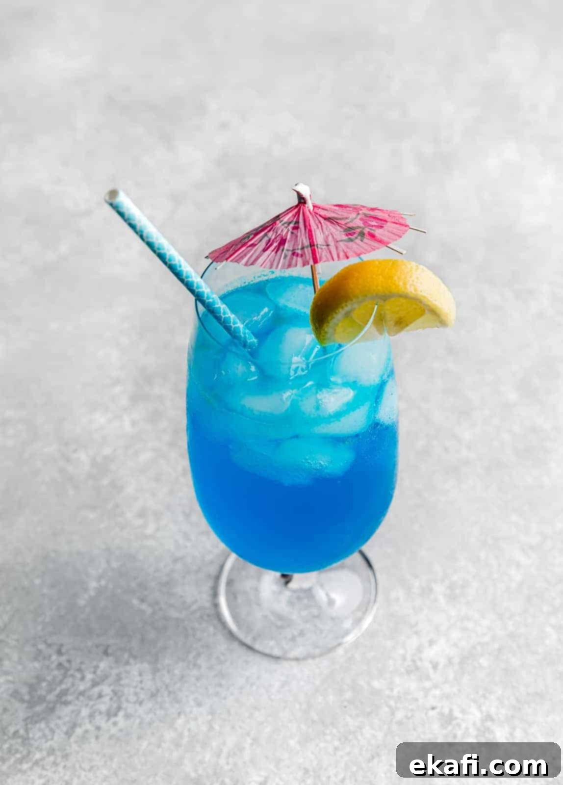 Γαλάζια Λιμνοθάλασσα Παρθένο Κοκτέιλ 3 Ένα Blue Lagoon mocktail σε γκρι φόντο με ροζ ομπρελίτσα και φέτα λεμονιού στο χείλος του ποτηριού.