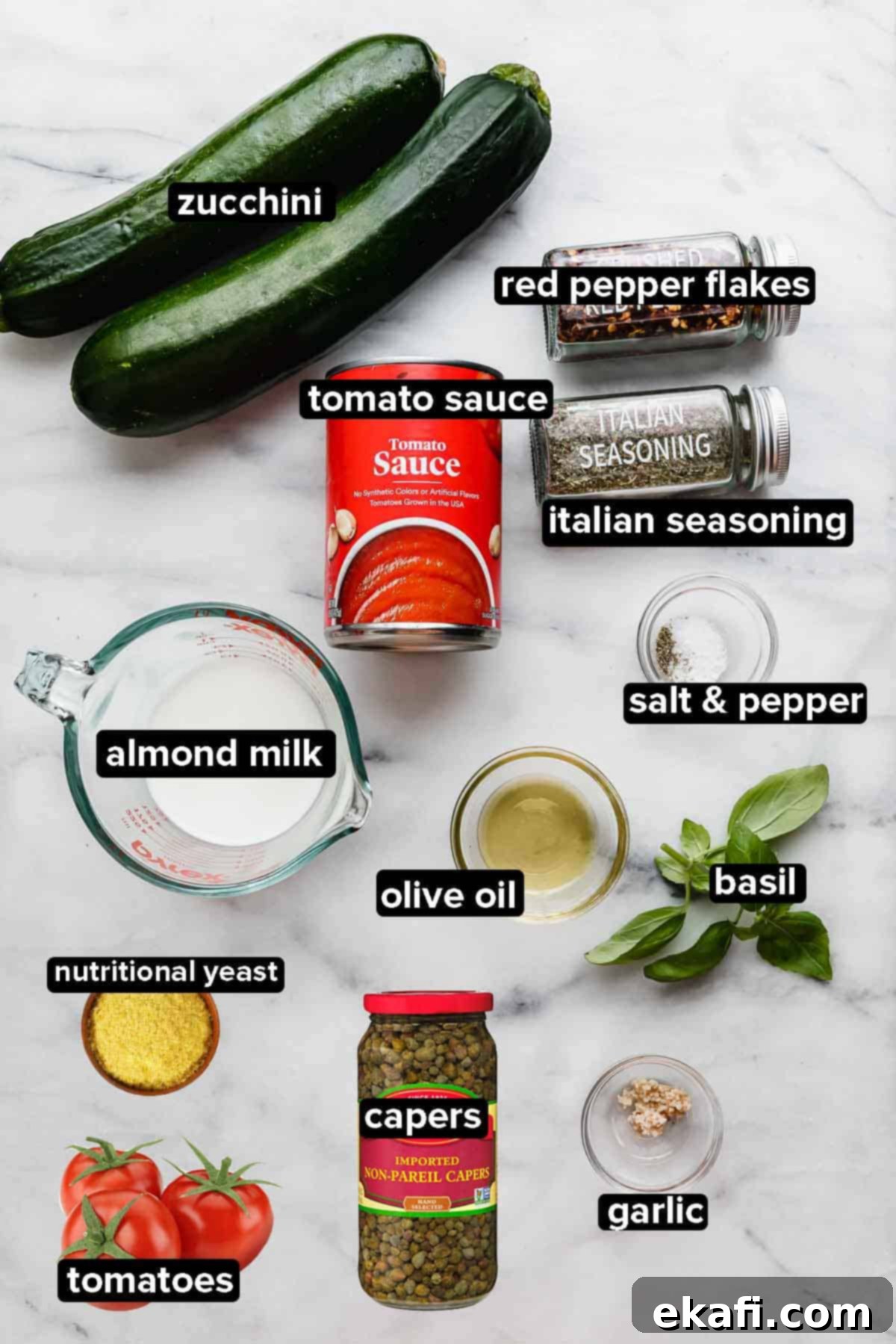 Fluted Zucchetti 3 Zucchetti Pasta ingredients on a white marble background, showcasing fresh zucchini, nutritional yeast, almond milk, ripe tomatoes, and Italian seasoning.