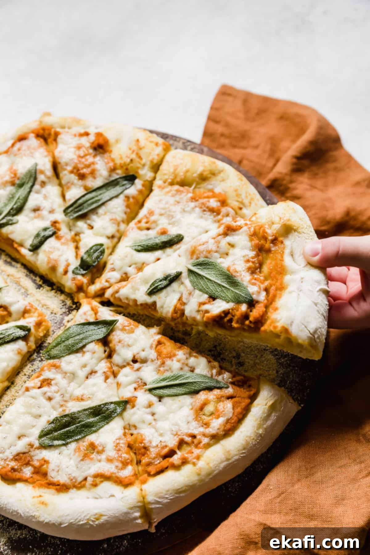 Harvest Moon Pizza 2 A hand pulling a slice of golden-crusted Pumpkin Pizza, topped with melted cheese and crispy sage, from the large pizza.