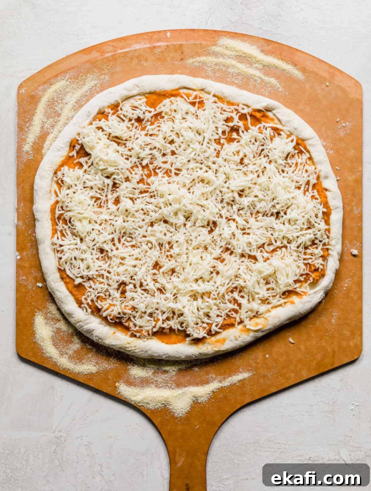 Harvest Moon Pizza 8 Shredded gouda cheese being sprinkled generously over a pumpkin puree topped pizza on a pizza peel, ready for the oven.