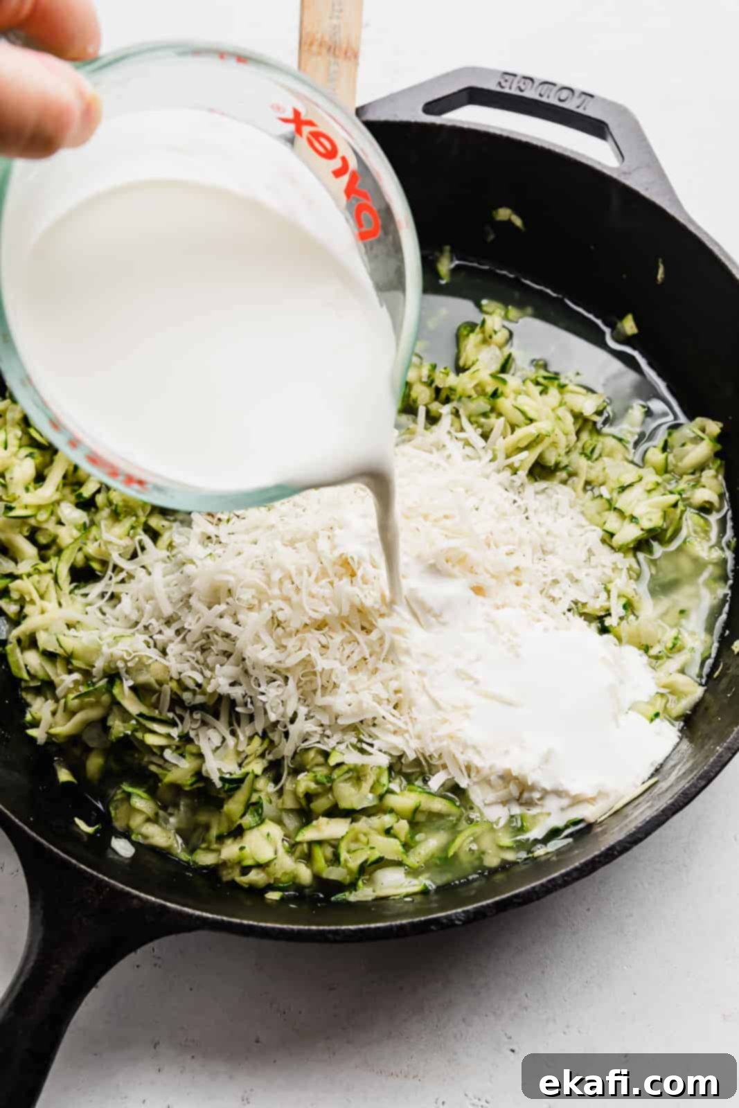 Summer Zucchini Delight 7 Heavy cream being poured overtop shredded zucchini that's in a black skillet.