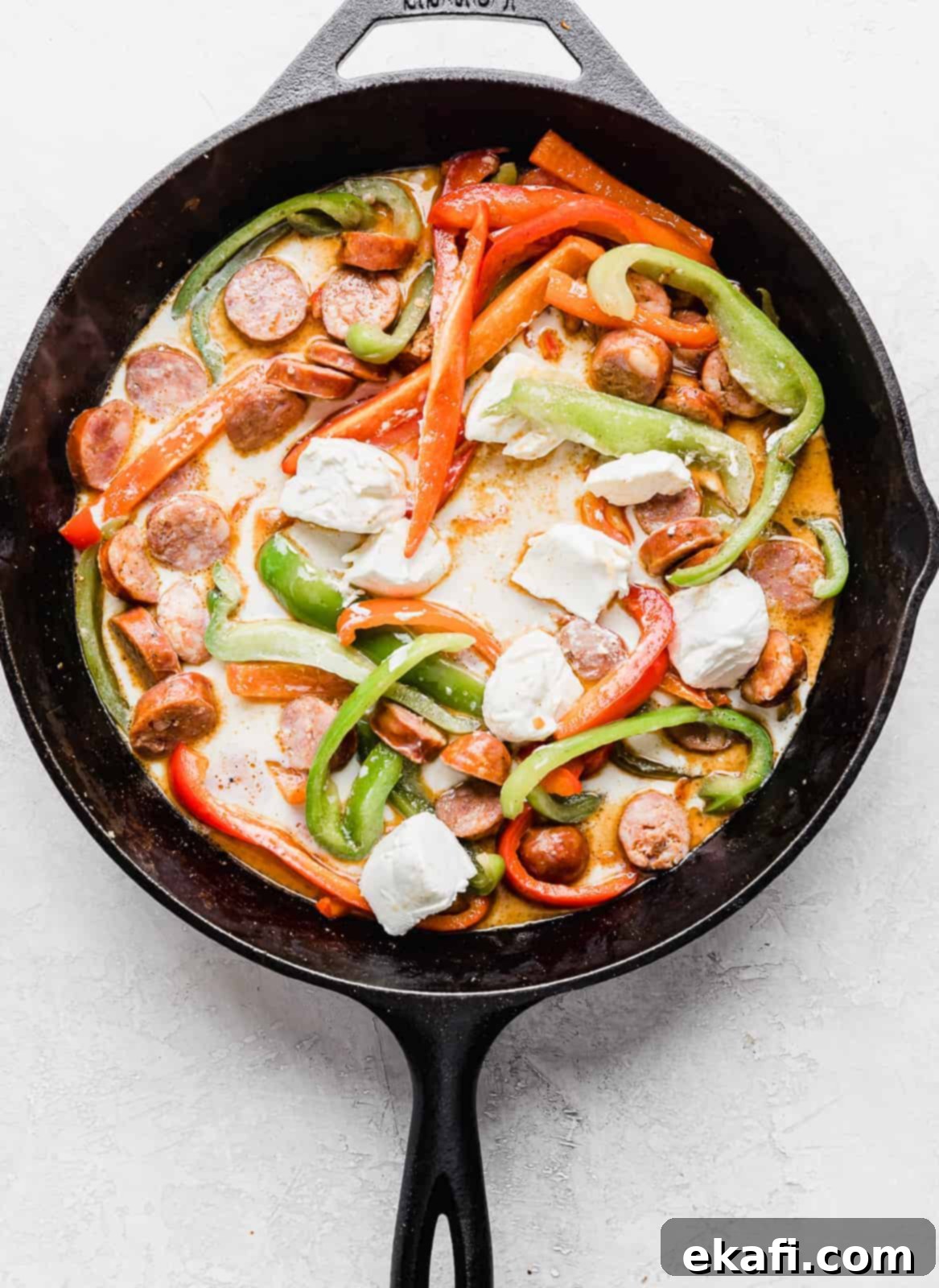 Spicy Cajun Sausage Pasta 6 A black skillet containing a colorful mix of sliced green and red peppers and sausage, with cubes of softened cream cheese beginning to melt into the ingredients, signifying the start of the creamy Cajun sauce for the pasta.