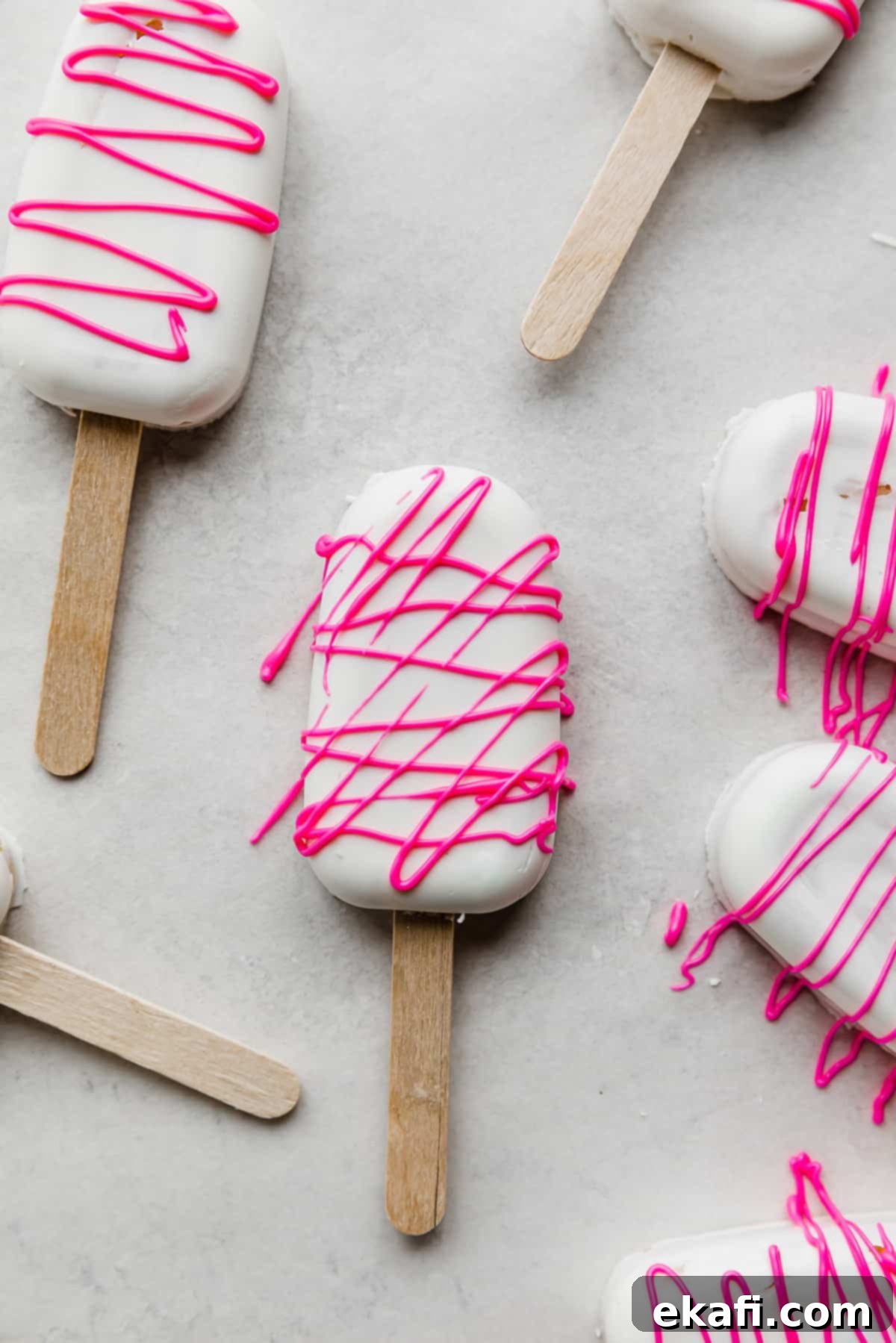 Κεκάκια Παγωτό 2 Λευκά Cakesicles καλυμμένα με σοκολάτα, με ροζ σοκολατένιο σιρόπι από πάνω, σε λευκό φόντο.