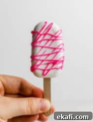 Κεκάκια Παγωτό 13 Ένα χέρι κρατά ένα Cakesicle καλυμμένο με λευκή σοκολάτα που έχει διακοσμηθεί με ροζ λιωμένη σοκολάτα.