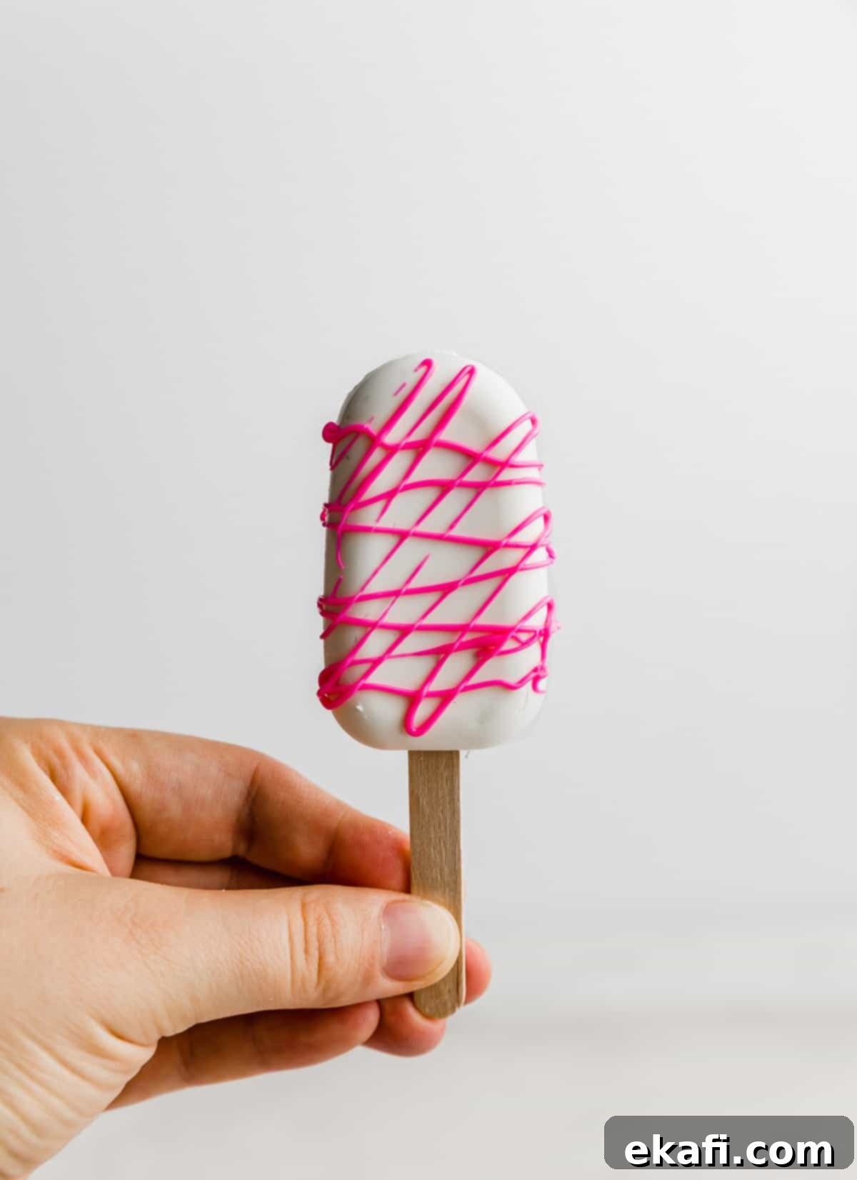 Κεκάκια Παγωτό 10 Ένα χέρι κρατά ένα Cakesicle καλυμμένο με λευκή σοκολάτα και ροζ σοκολατένιο σιρόπι.