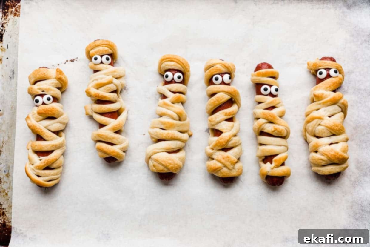 Mummy Hot Dogs 8 Mummy Hot Dogs on a white parchment lined baking sheet, with candy eyes on the mummy dogs.