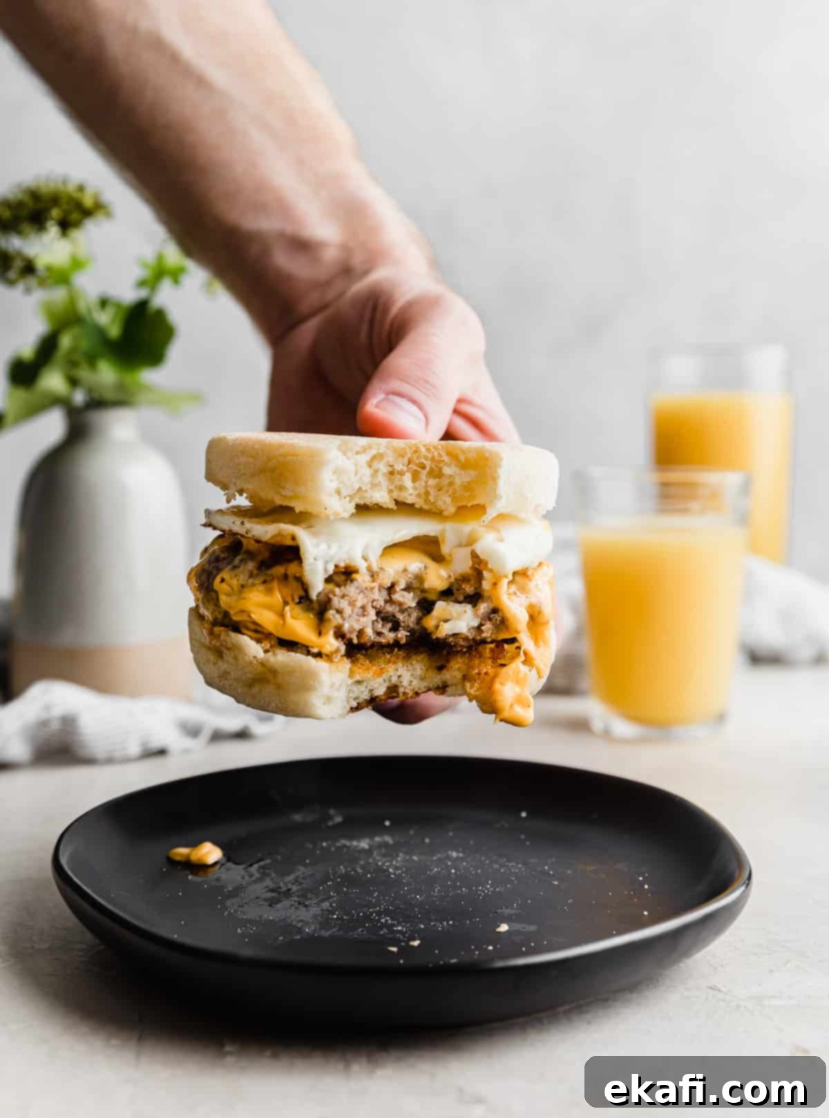 From Scratch Sausage and Egg Breakfast Sandwich 11 A hand holding a Homemade Sausage and Egg McMuffin with a generous bite taken out, revealing the layers of fluffy egg, melted cheese, and seasoned sausage patty, against a gray background.