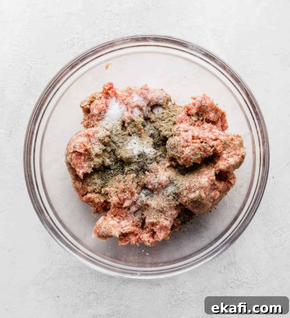 From Scratch Sausage and Egg Breakfast Sandwich 4 A clear glass bowl containing ground pork mixed with various dry seasonings like sage, thyme, onion powder, salt, and pepper, ready to be formed into patties.