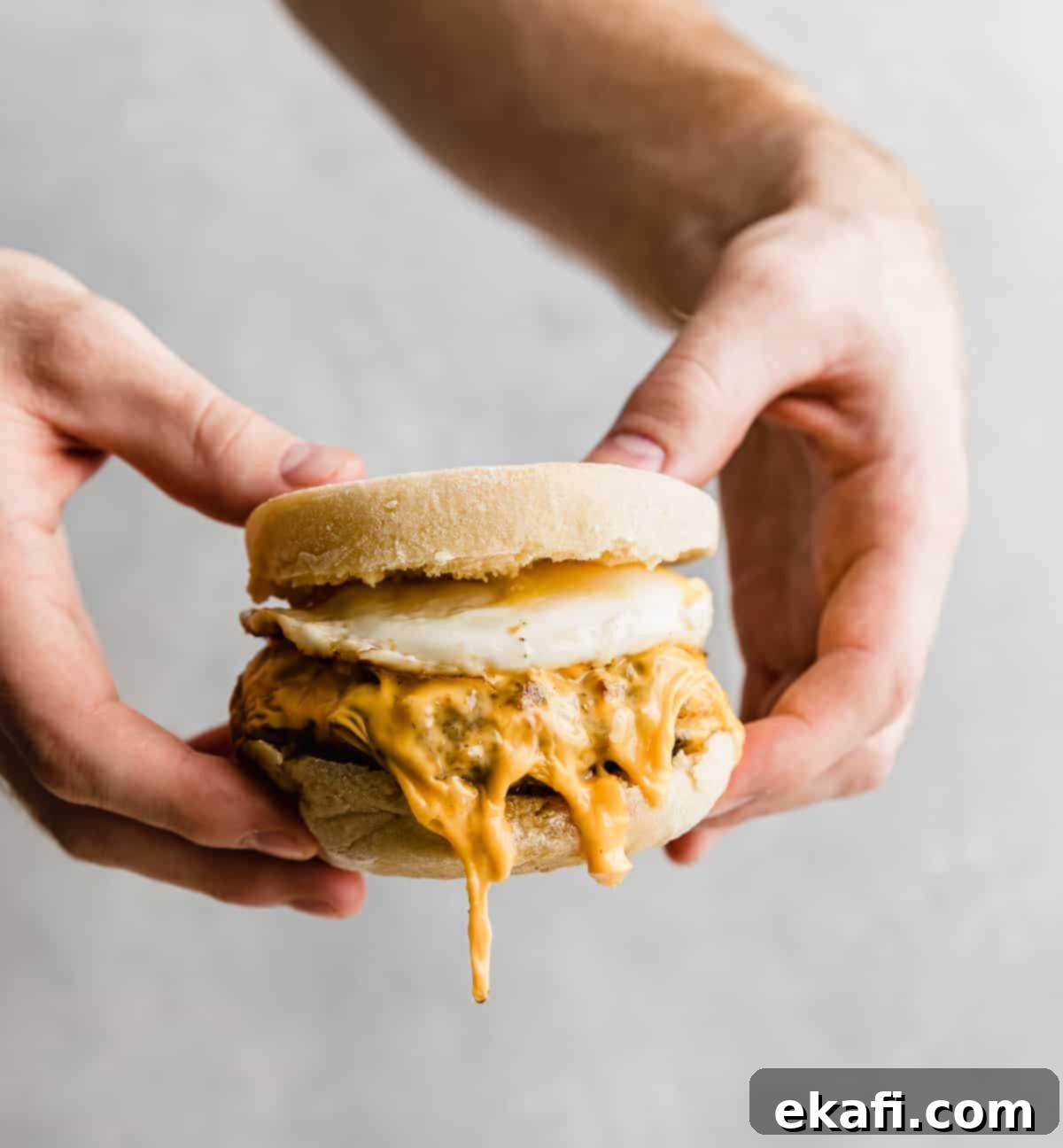 From Scratch Sausage and Egg Breakfast Sandwich 10 A set of hands carefully holding a fully assembled Homemade Sausage and Egg McMuffin against a soft gray background, ready to be enjoyed.