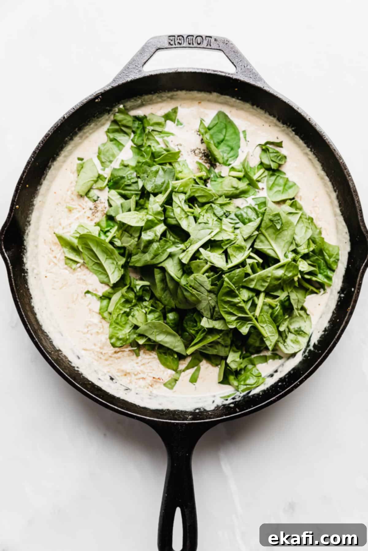 Tuscan Garlic Cream Chicken 5 Spinach overtop a tan tuscan chicken gravy mixture in a skillet on a white background.