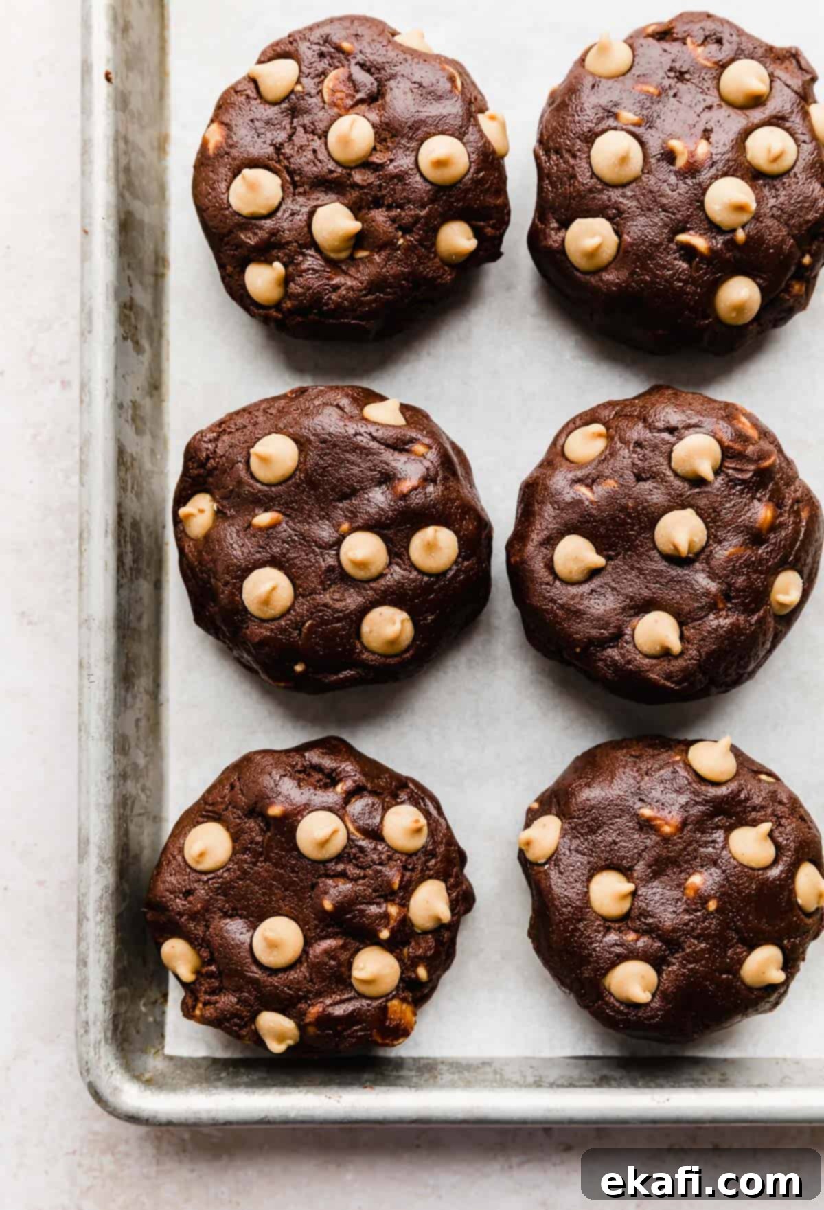Decadent Peanut Butter Chocolate Chip Cookies 5 Large, unbaked Chocolate Peanut Butter Chip Cookie dough balls, flattened into puck shapes, neatly arranged on a white parchment-lined baking sheet.