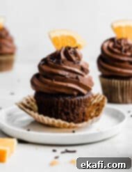 Zesty Chocolate Orange Delights 14 A chocolate frosting topped chocolate orange cupcake on a white plate, with a fresh slice of an orange as a topper.