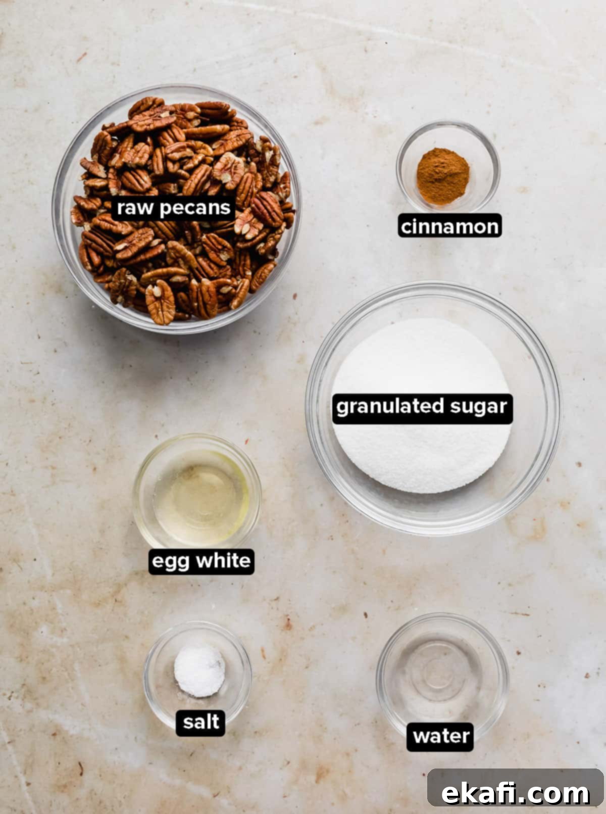 Sweet Glazed Pecans 3 Collection of ingredients for candied pecans, including raw pecans, sugar, cinnamon, salt, and an egg white, displayed in glass bowls on a marble background.