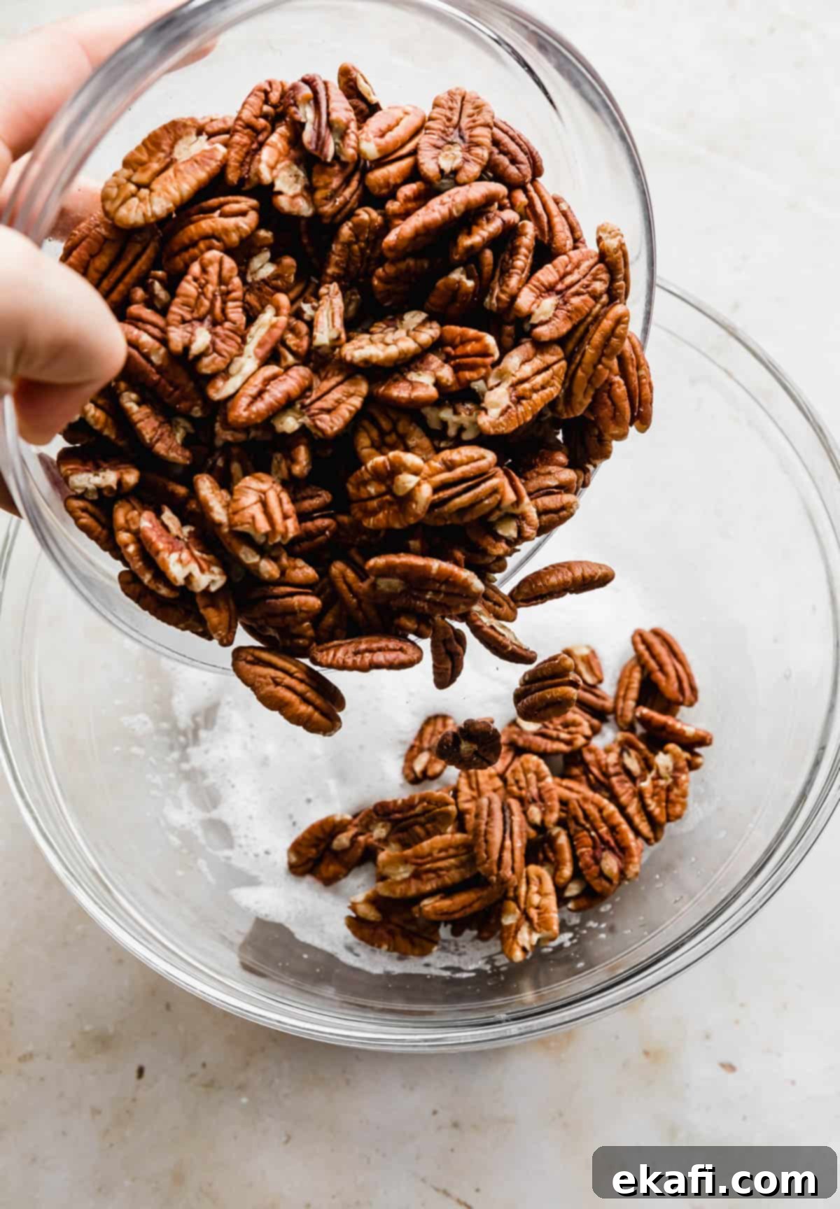 Sweet Glazed Pecans 6 Raw pecan halves being gently poured into the frothy egg white mixture in a glass bowl.