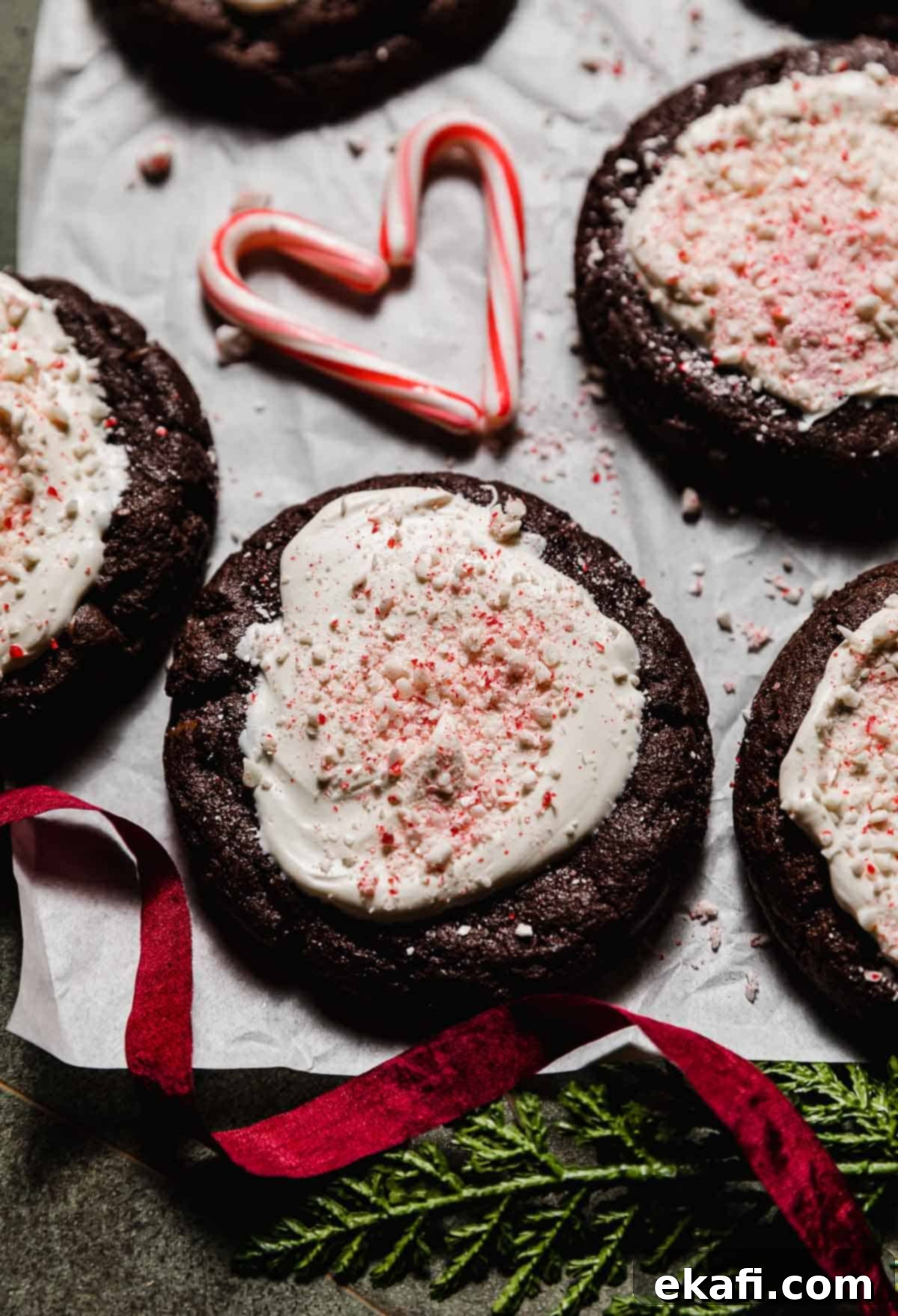 Chocolate Peppermint Bark Cookies 8 Chocolate cookie topped with white chocolate and crushed candy canes (Peppermint Bark Cookie) on a white parchment paper with a small candy cane heart in the background.