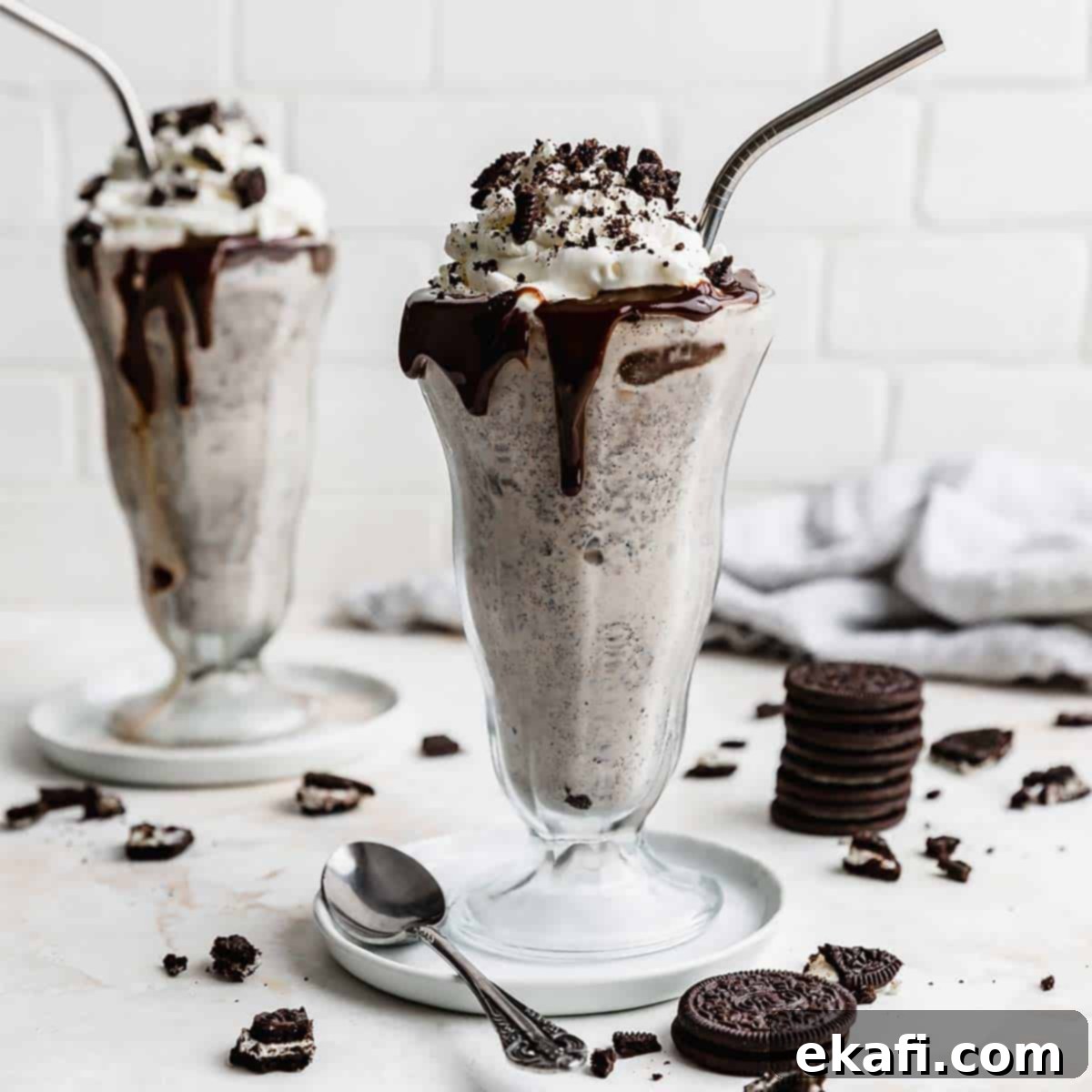 Irresistible Oreo Desserts 3 Two milkshake glasses filled with the best Oreo Milkshake recipe, against a white brick background with Oreo crumbs scattered around the cups.