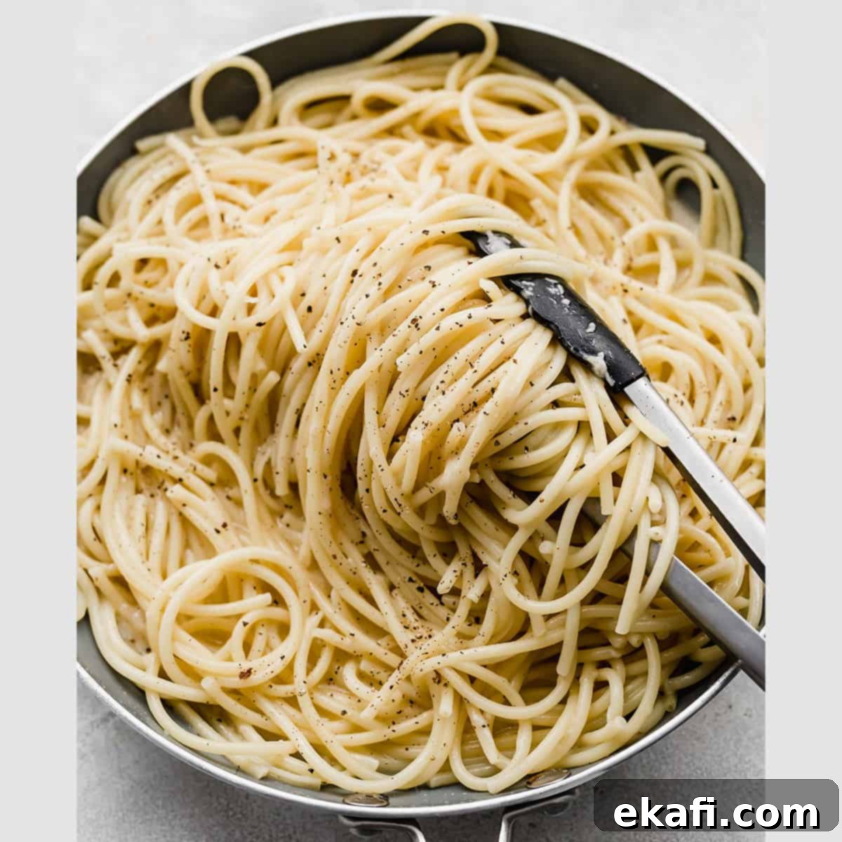 Το Παρασκευιάτικο Τραπέζι 8 Bucatini Cacio e Pepe σε τηγάνι με λαβίδα κουζίνας να γυρίζει τα ζυμαρικά.