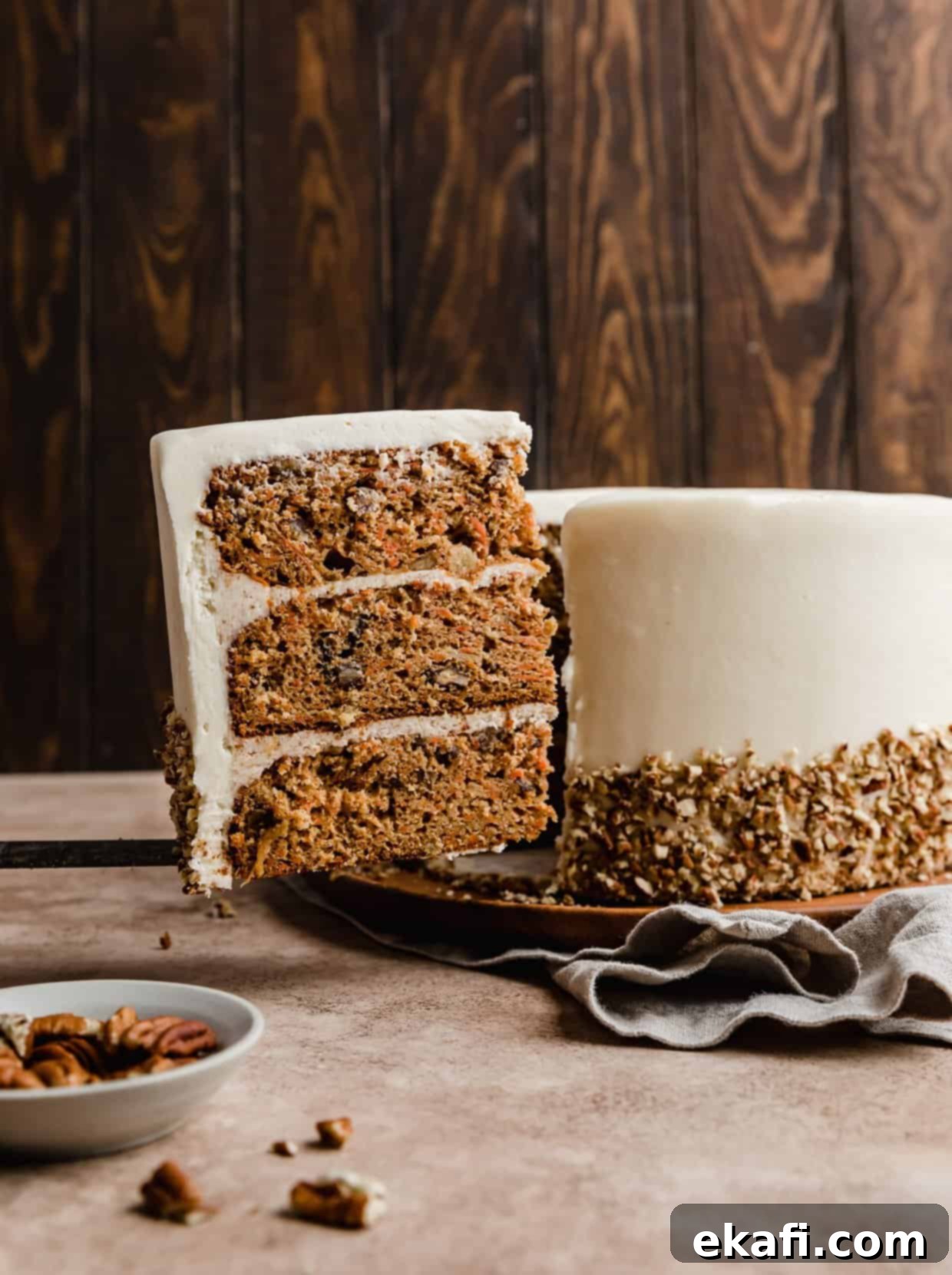Tropical Pecan Carrot Cake 2 A perfectly sliced piece of layered Carrot Cake with Pineapple and Pecans, showcasing the rich cream cheese frosting and delightful filling, set against a warm brown background. The entire frosted carrot cake is visible in the soft background blur.