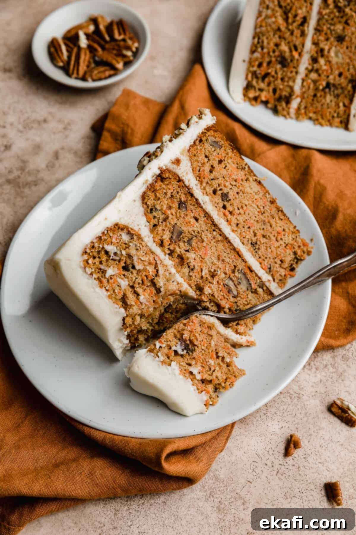 Tropical Pecan Carrot Cake 3 A beautifully frosted slice of carrot cake with visible pecans and pineapple chunks, presented on a pristine white plate, inviting a taste of its moist texture.