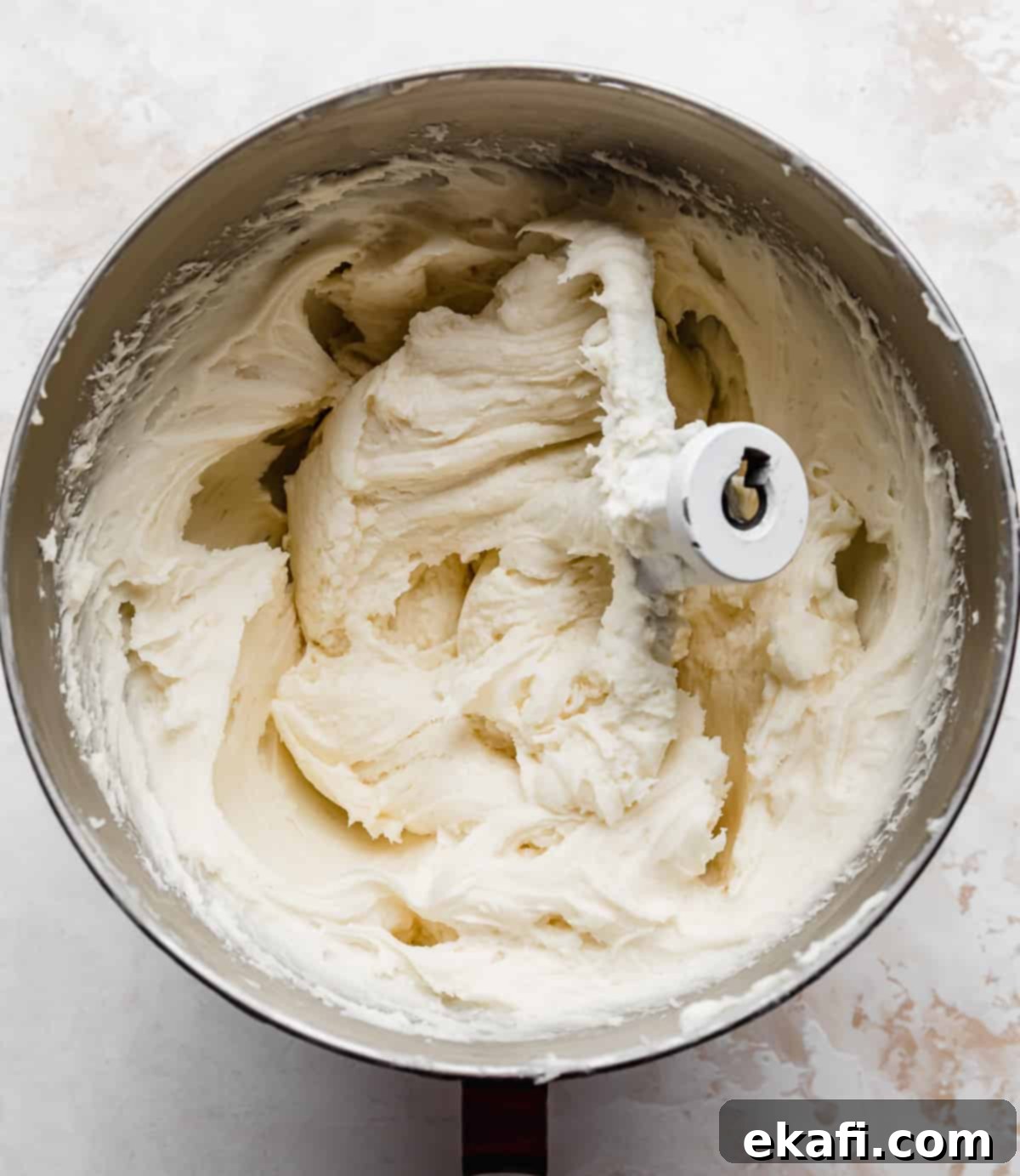 Tropical Pecan Carrot Cake 4 A large metal mixing bowl filled with freshly prepared, smooth and creamy white cream cheese frosting, ready for decorating, set against a clean white background.