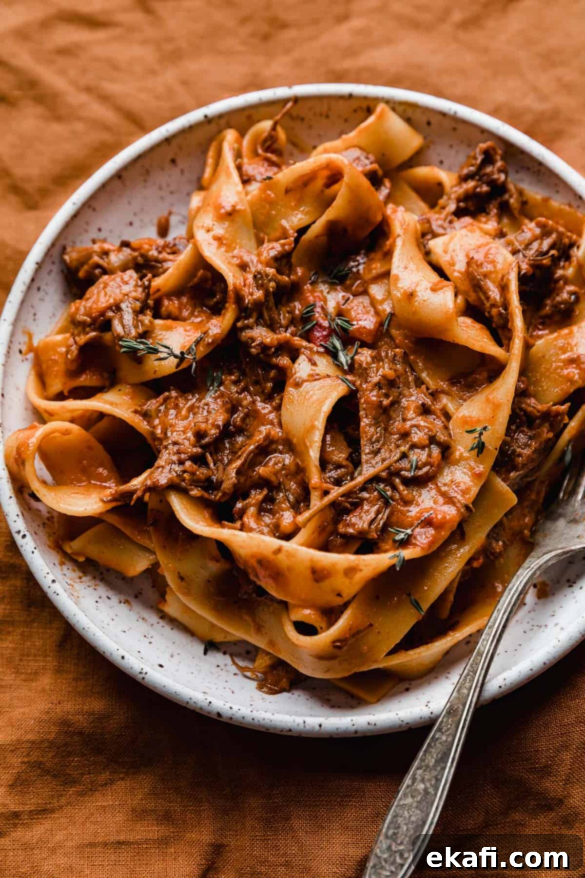 Hearty Shredded Beef Ragu 2 A white plate with the best beef Ragu made with pappardelle noodles, on a burnt orange background.