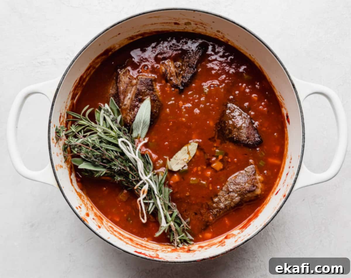 Hearty Shredded Beef Ragu 7 A white pot with fresh herbs bundled with kitchen twin a top beef chuck roast in a beef Ragu sauce.