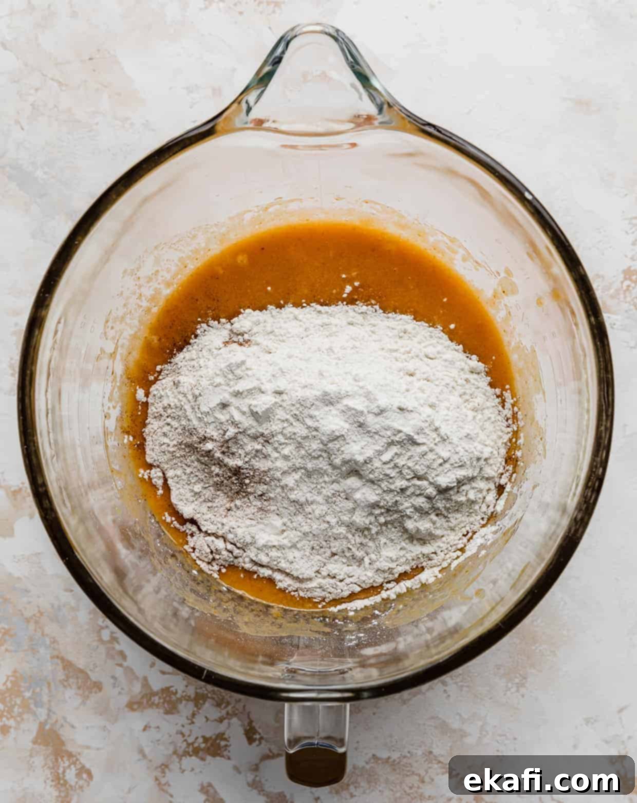 Spiced Pumpkin Chocolate Chip Delights 10 A layer of flour ingredients gently sifted over an orange liquid mixture in a clear glass mixing bowl, initiating the combination for pumpkin chocolate chip cookies.
