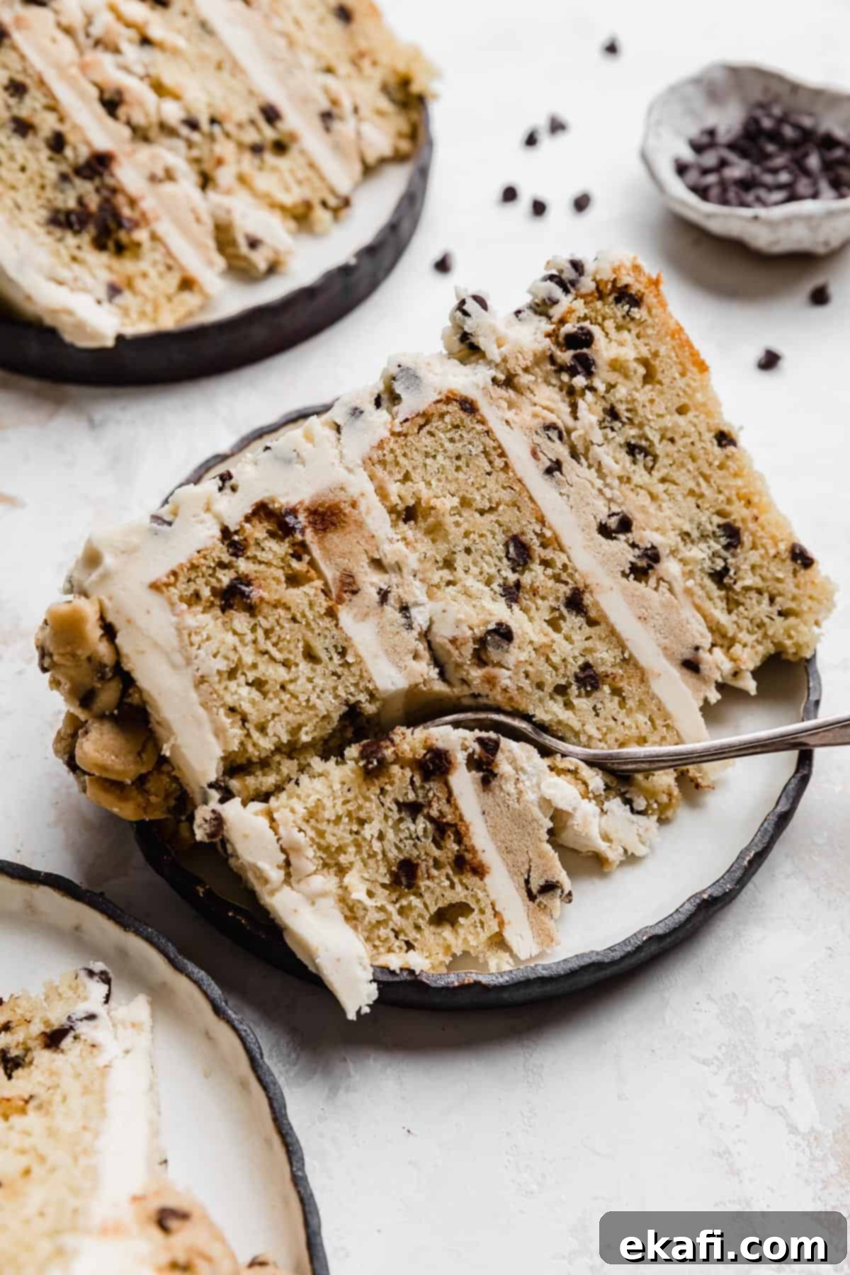 The Ultimate Cookie Dough Dream Cake 2 A beautifully presented slice of Brown Butter Cookie Dough Cake on a white plate, revealing its distinct layers of cake, edible cookie dough, and creamy frosting. A fork is cutting into the top.