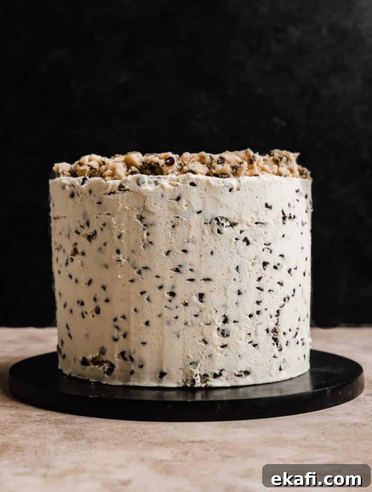 The Ultimate Cookie Dough Dream Cake 10 A finished three-layered round Cookie Dough Cake frosted with white frosting and mini chocolate chips, set against a dark background.