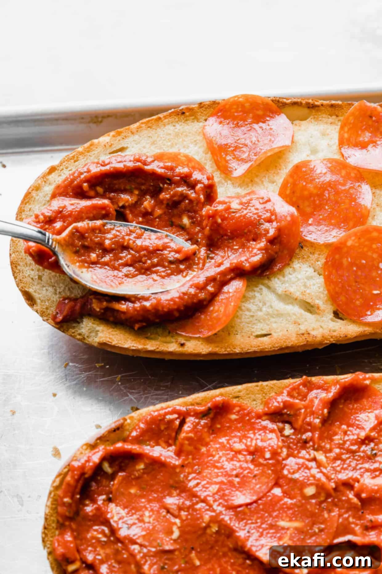 French Bread Pizza 8 A spoon spreading pizza sauce overtop of pepperonis on a French bread loaf.